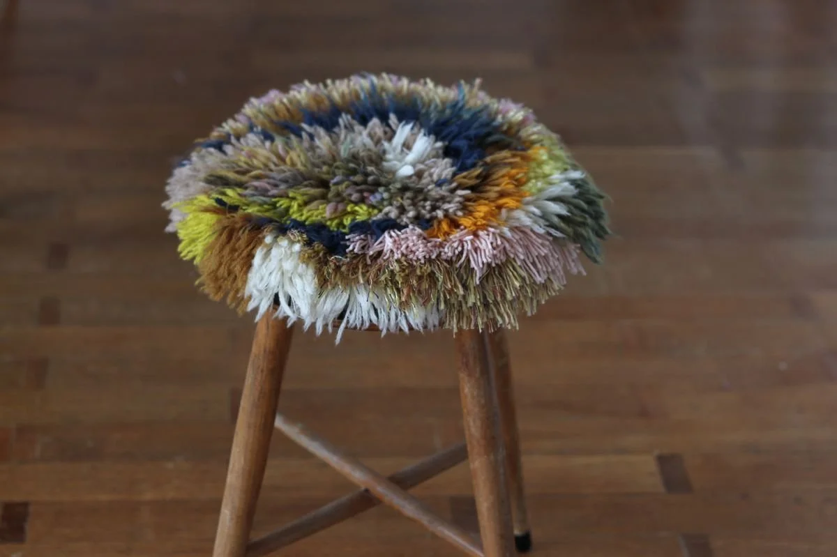 A small wooden stool with a colorful, textured, shaggy cushion on top, placed on a hardwood floor.