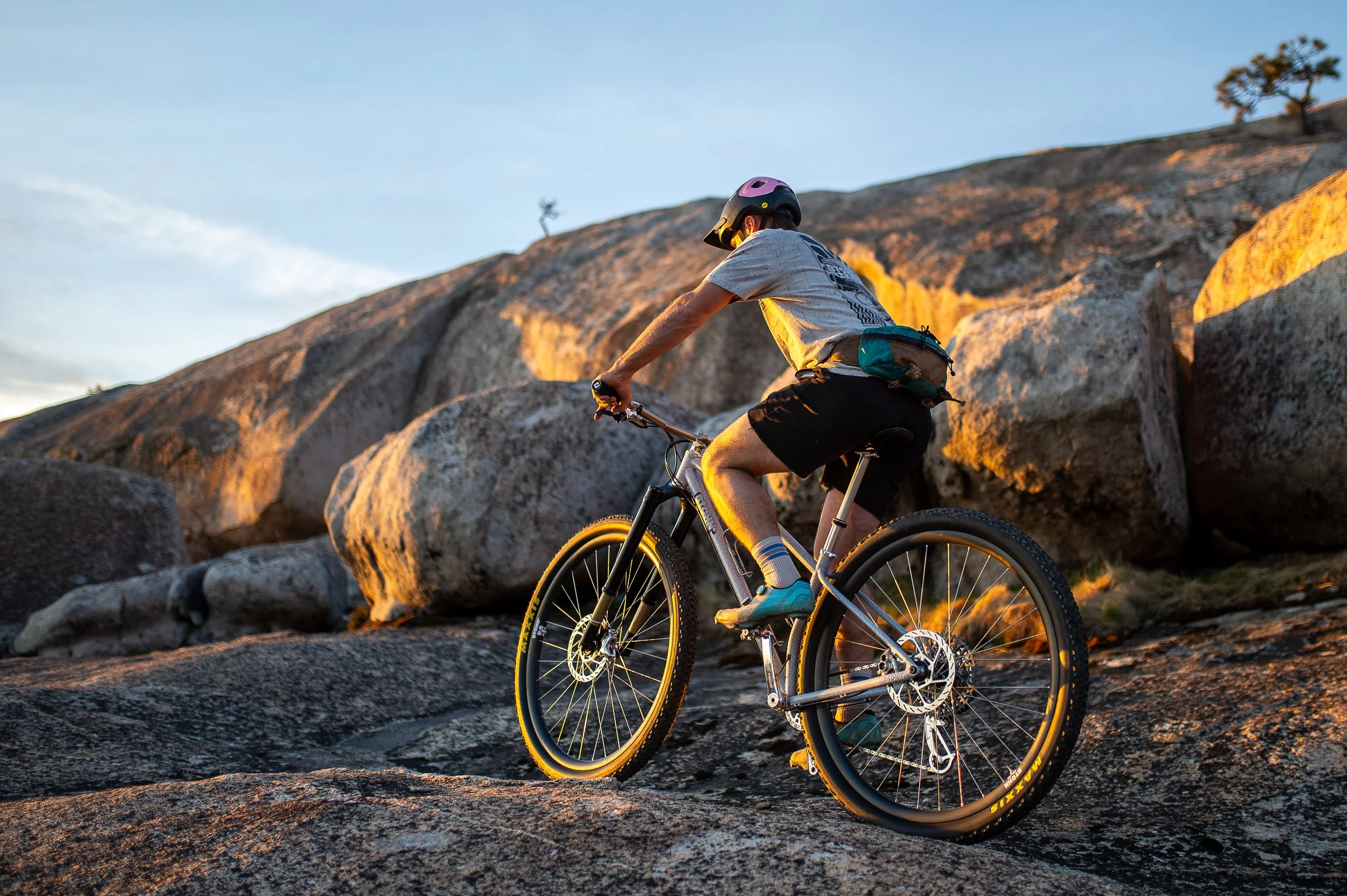 A person riding a mountain bike on rocky terrain during sunset, wearing a helmet, t-shirt, shorts, and sneakers.