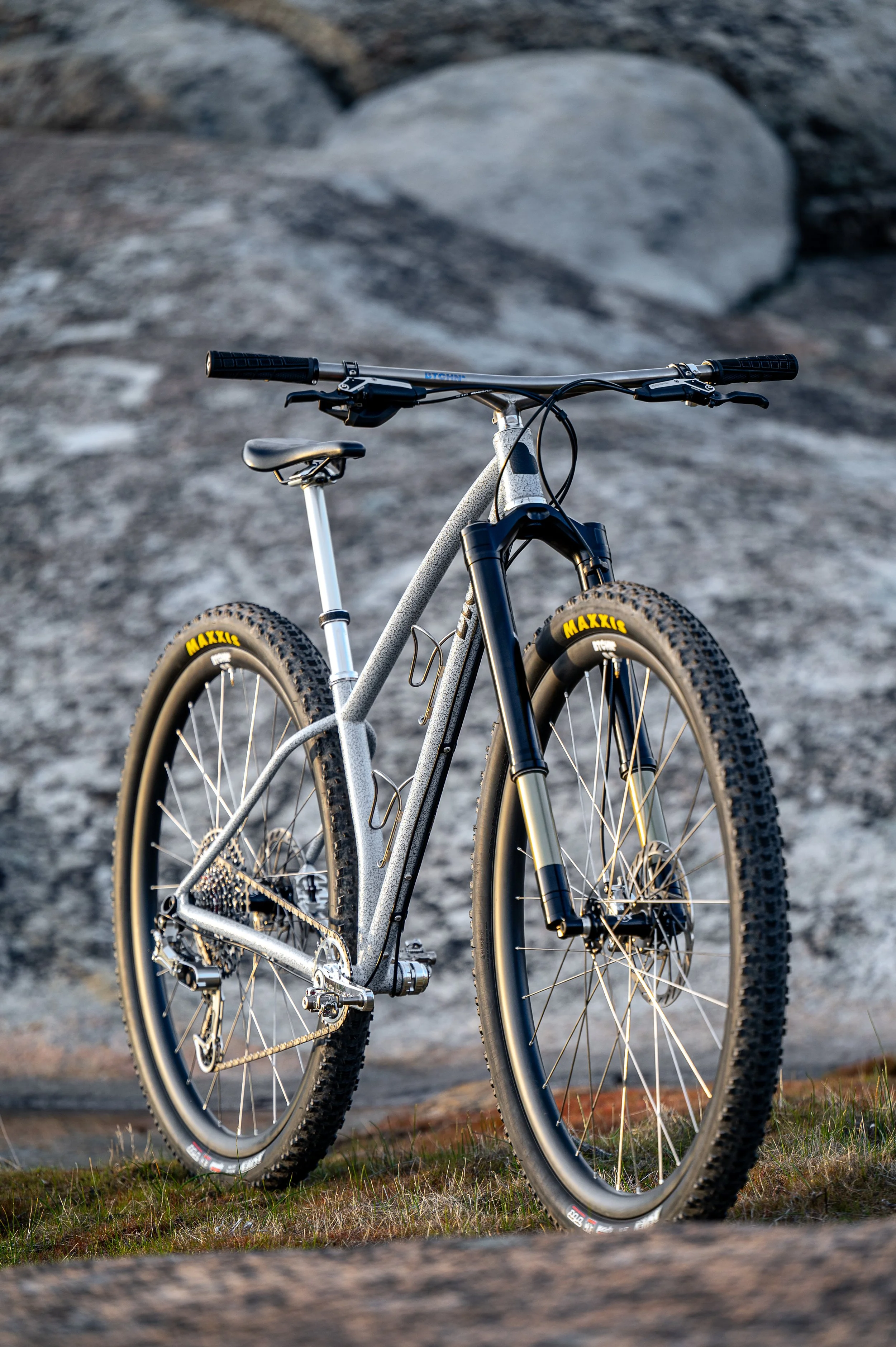 A mountain bike with large tires and a sleek frame, resting on grassy terrain with rocks in the background.