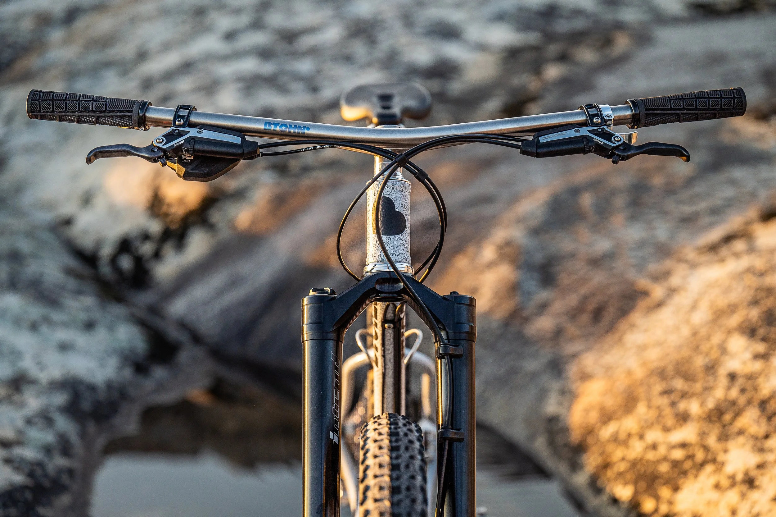 Front view of a mountain bike with a black suspension fork and wide knobby tires, positioned on a rocky trail at sunset.