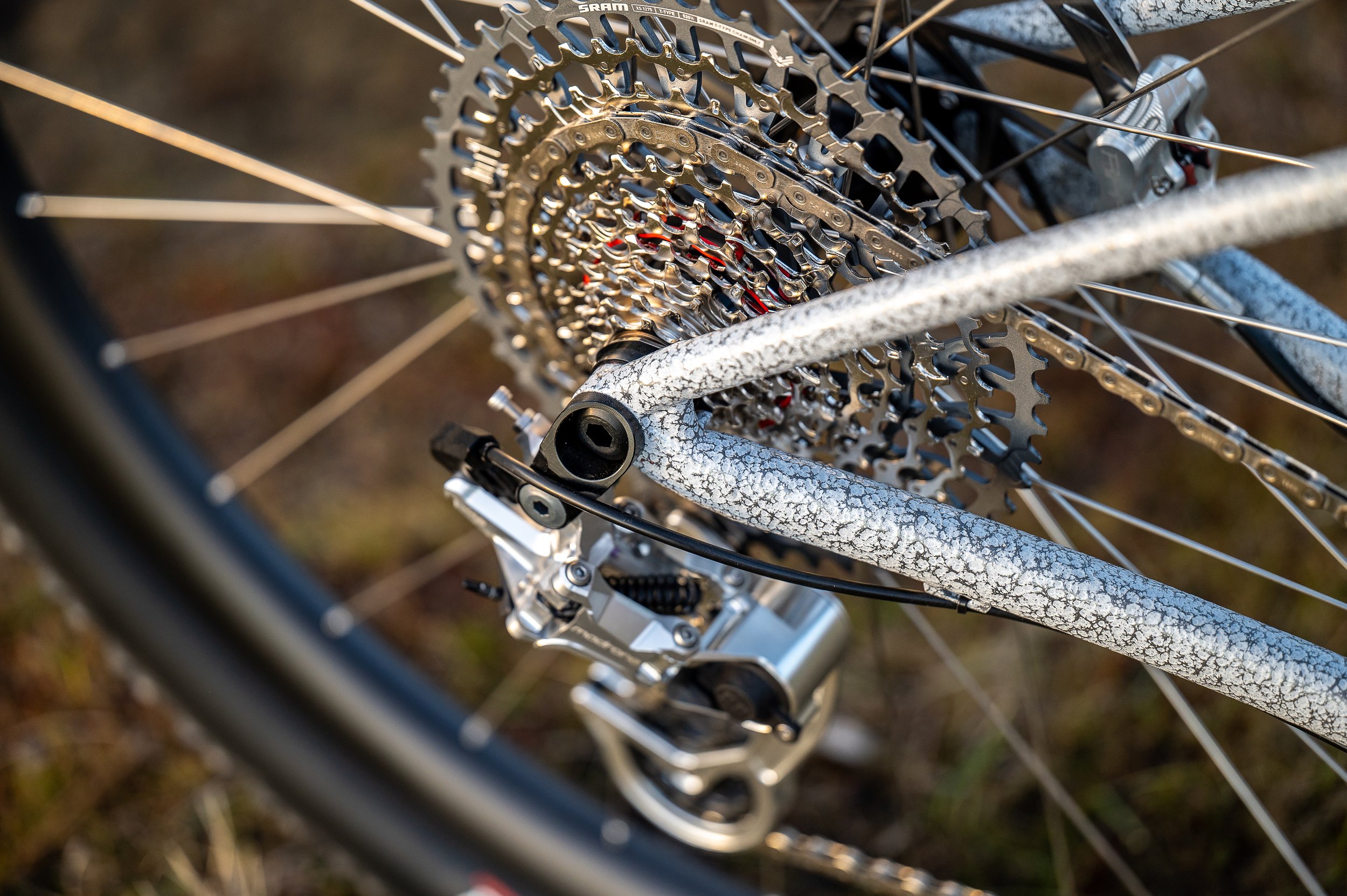 Close-up of a mountain bike's rear gear cassette, derailleur, and frame with a gravel texture, set against a blurred natural background.