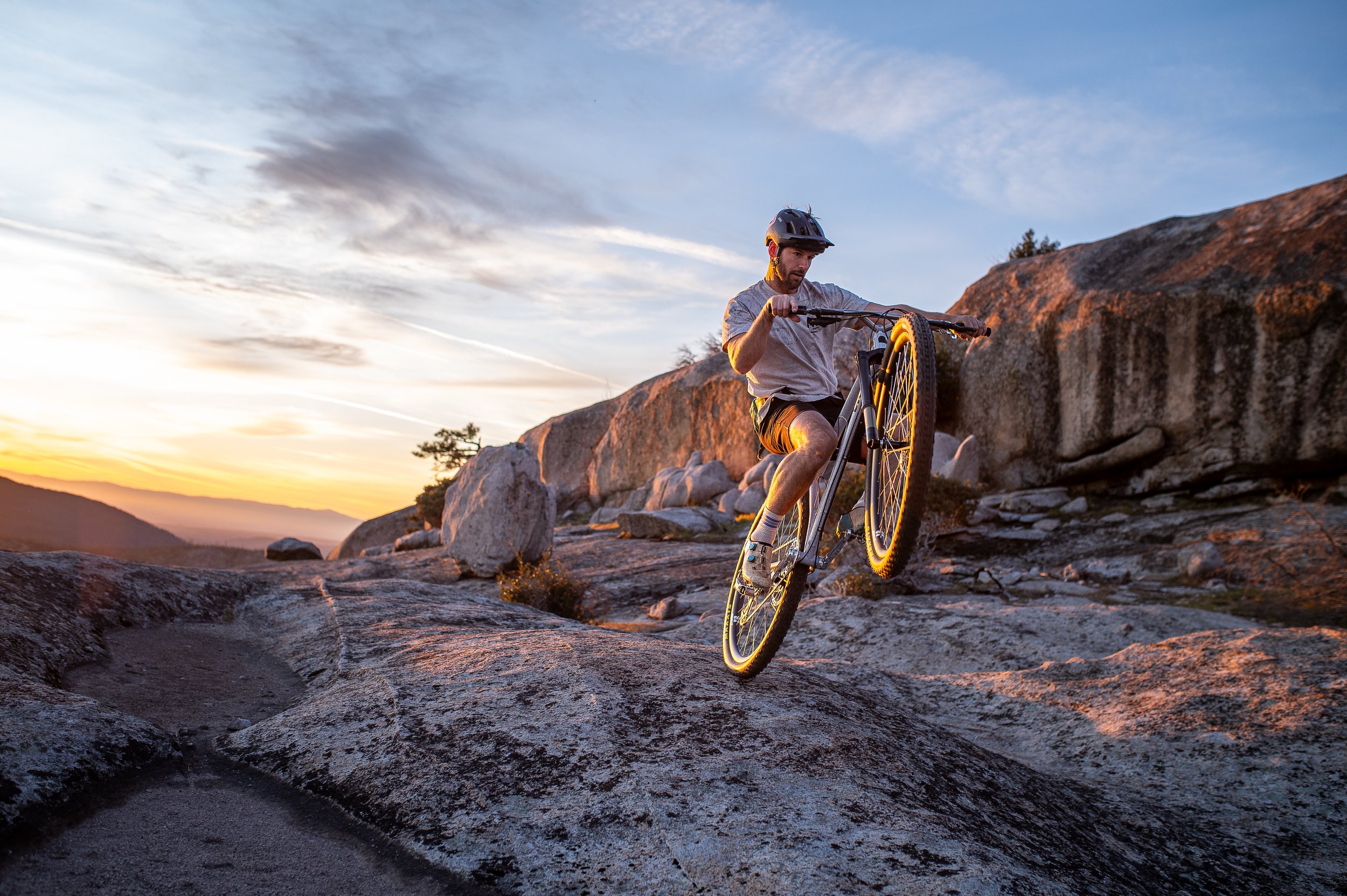 A man riding a mountain bike on rocky terrain during sunset, wearing a helmet and casual clothing.
