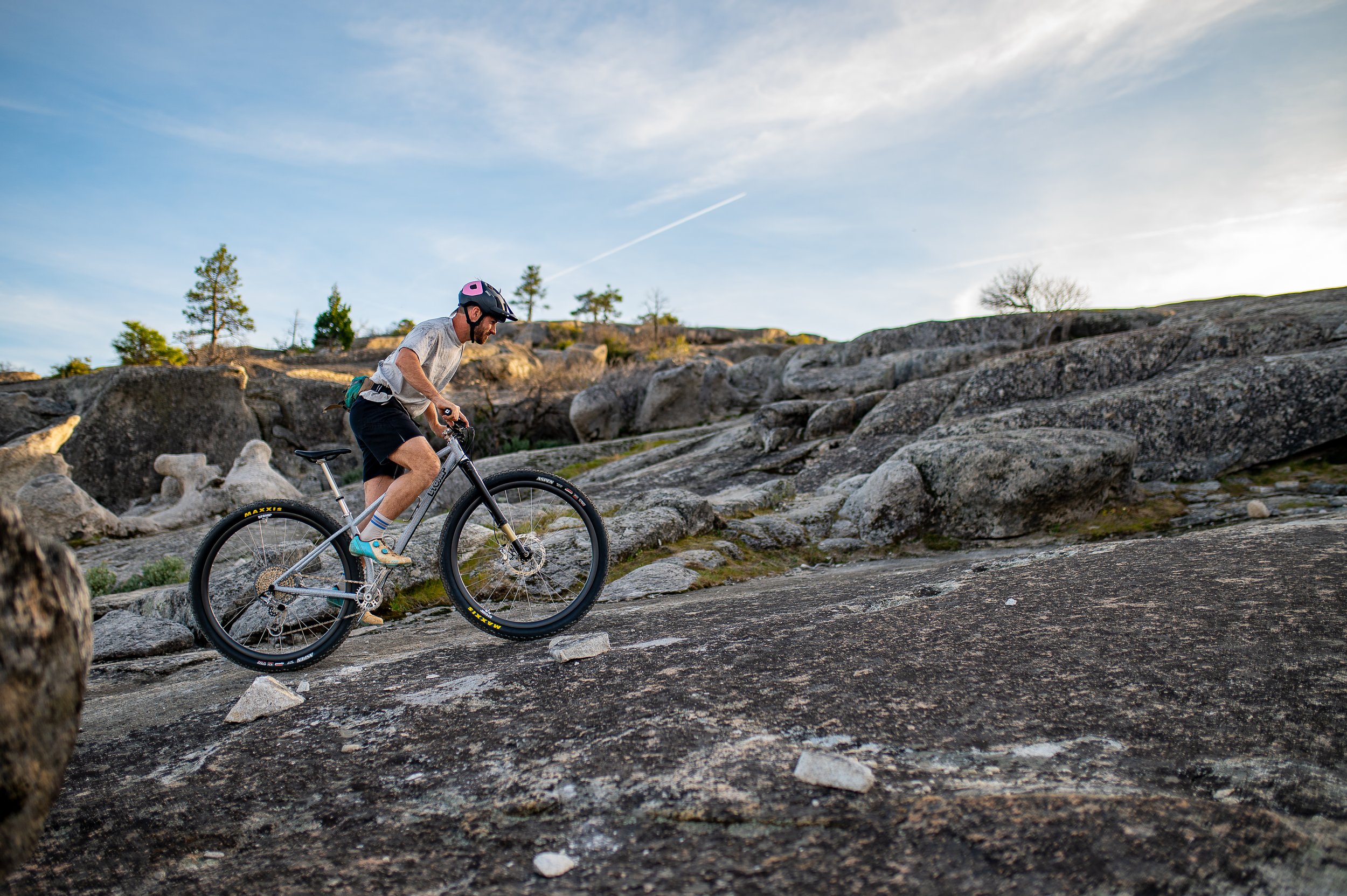 A person riding a mountain bike on a rocky incline outdoors with trees and a blue sky in the background.