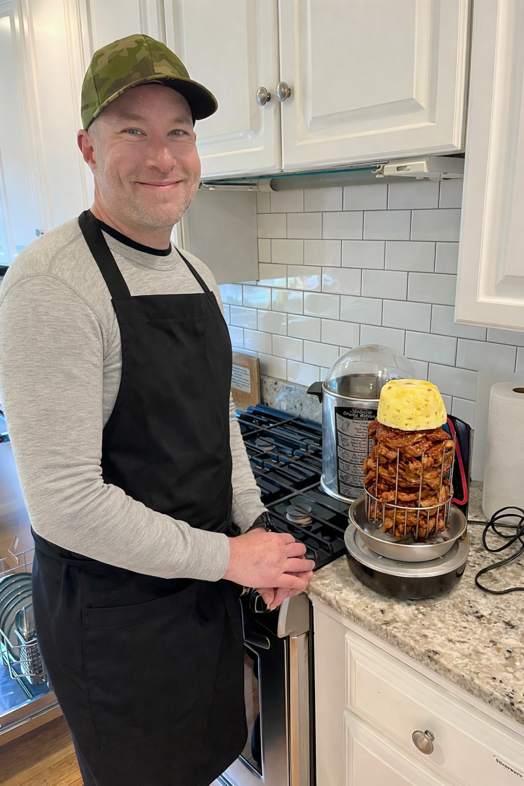 A man wearing a black apron is standing in a kitchen next to a countertop. On the counter, there is cooked chicken stacked vertically, topped with a yellow cheese block, and a device heating the chicken. The man is smiling at the camera.