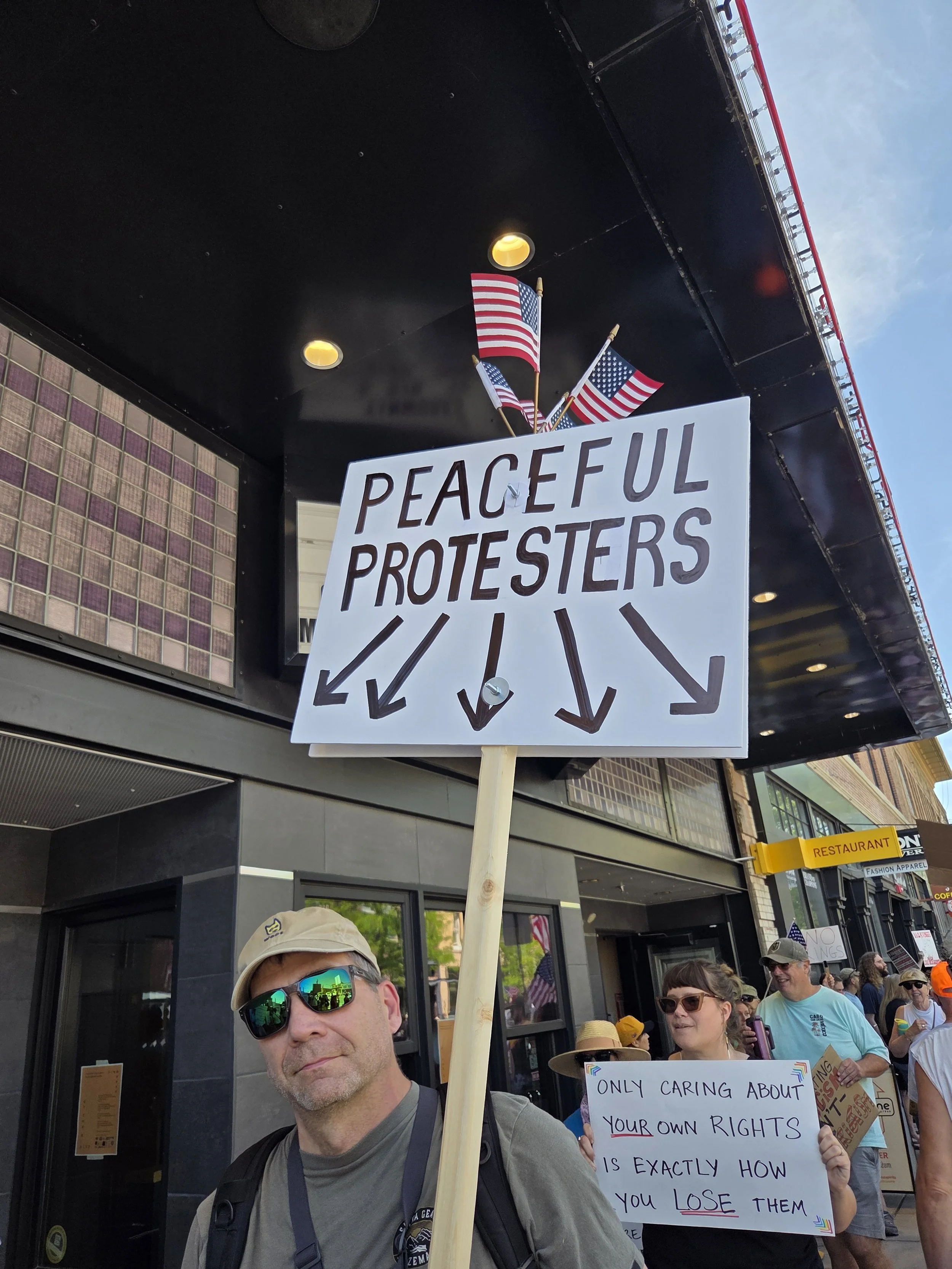 Protester holding a sign that reads 'Peaceful Protesters' with arrows pointing down, surrounded by other protesters, some holding signs, on a city street with American flags hanging above.