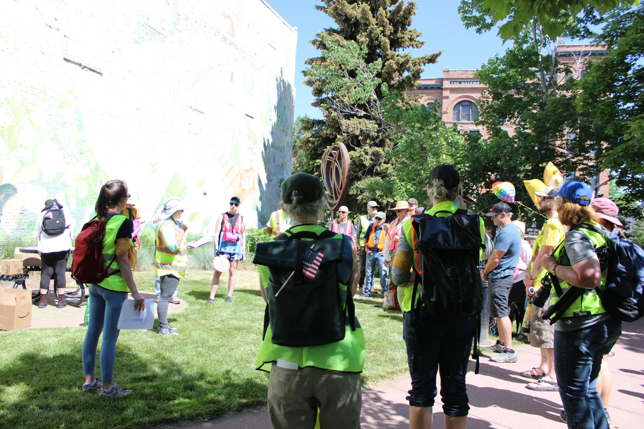 A group of people wearing yellow safety vests and backpacks gathered outdoors around a speaker, in a park surrounded by trees and a mural on a wall.