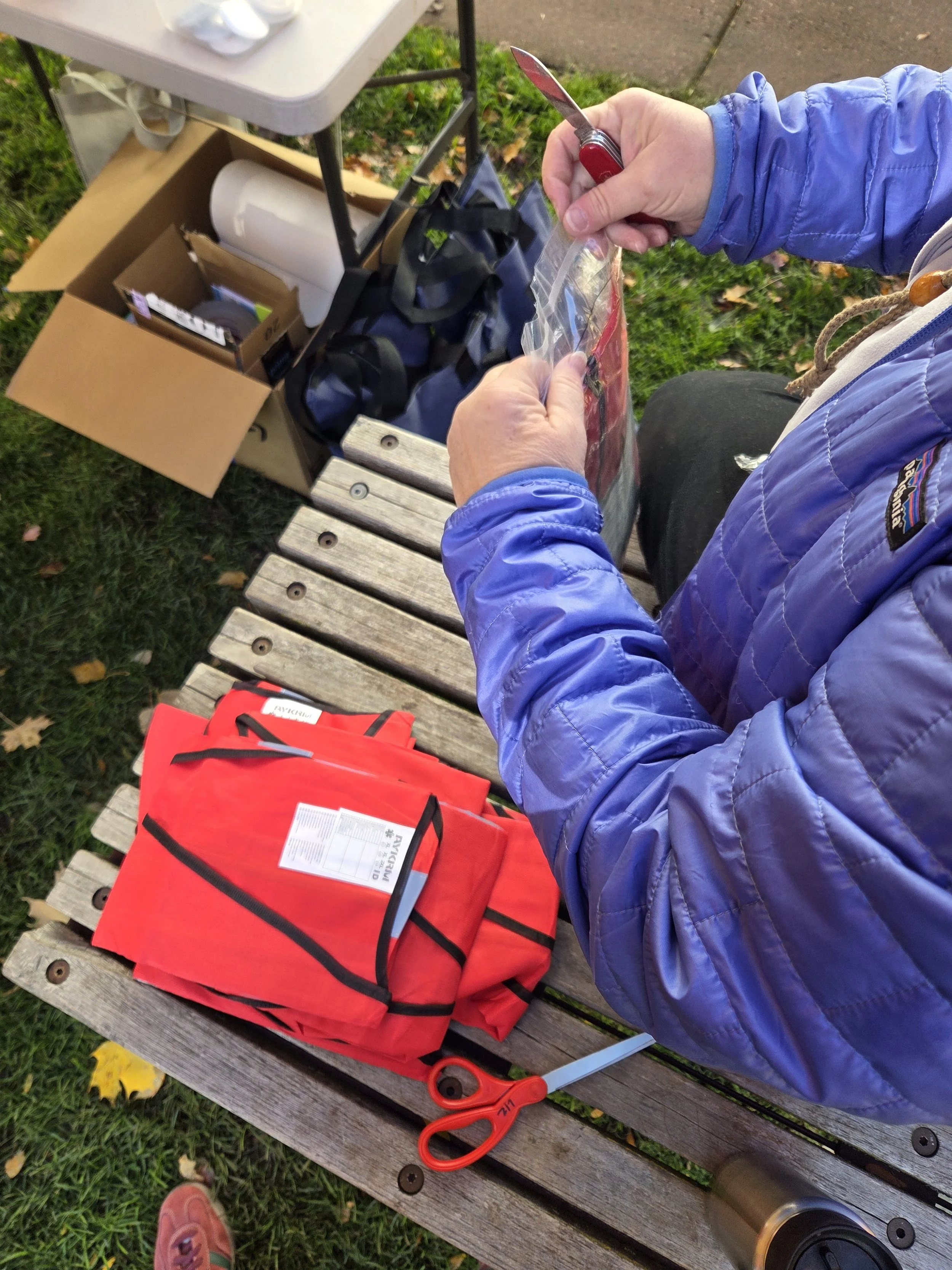 A person wearing a blue jacket is using scissors to cut a clear plastic bag while sitting on a wooden bench outdoors. Nearby on the bench are a folded red life vest, a pair of scissors, and a white tag. There is a cardboard box and a black bag on a small table behind the person, with some items inside, including a white roll of paper.