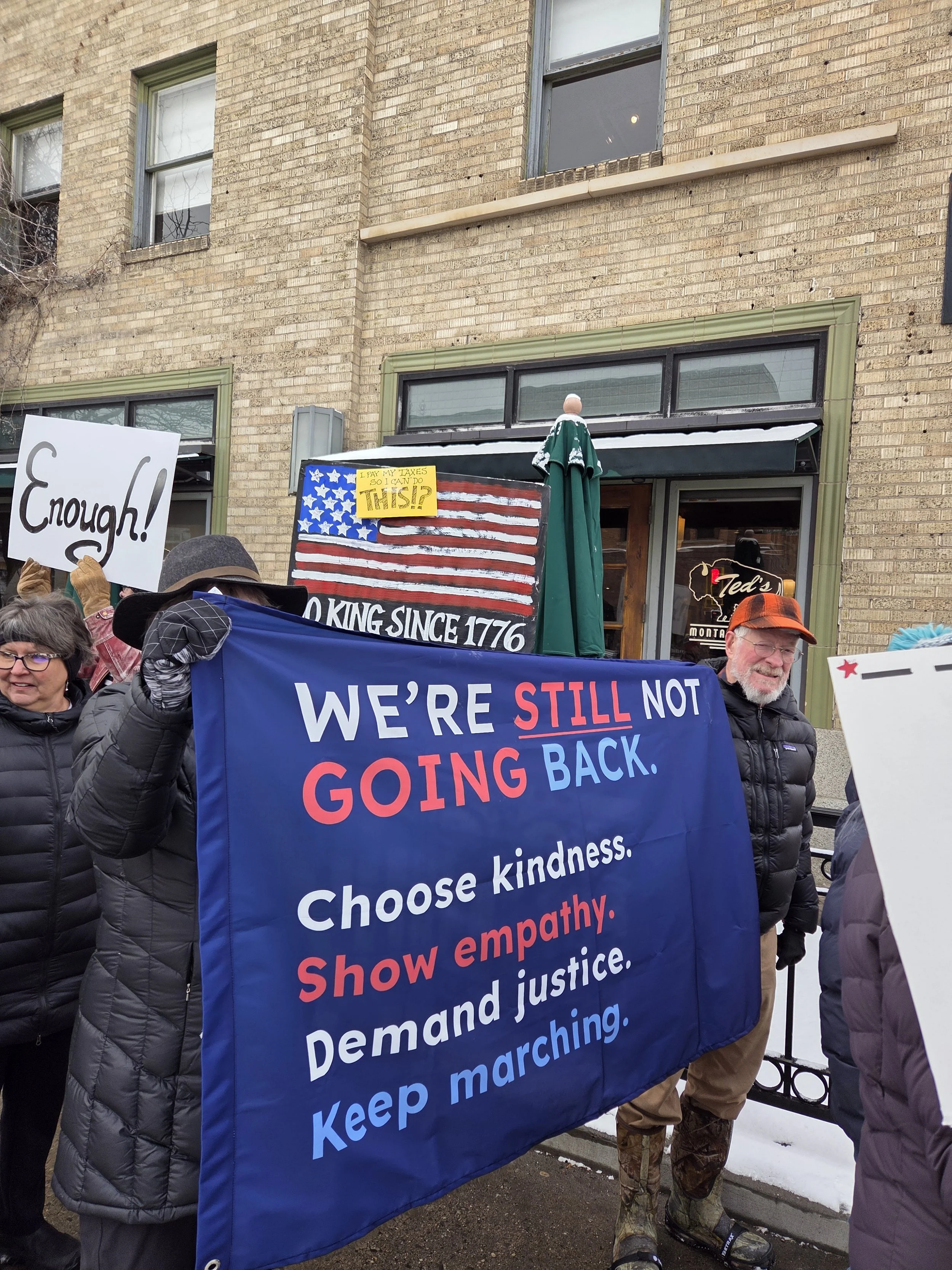 Protesters holding a large blue banner with messages: "We're still not going back. Choose kindness. Show empathy. Demand justice. Keep marching." Others hold signs, one says "Enough!" and another reads "Pay my taxes so I can do this!" The group is st