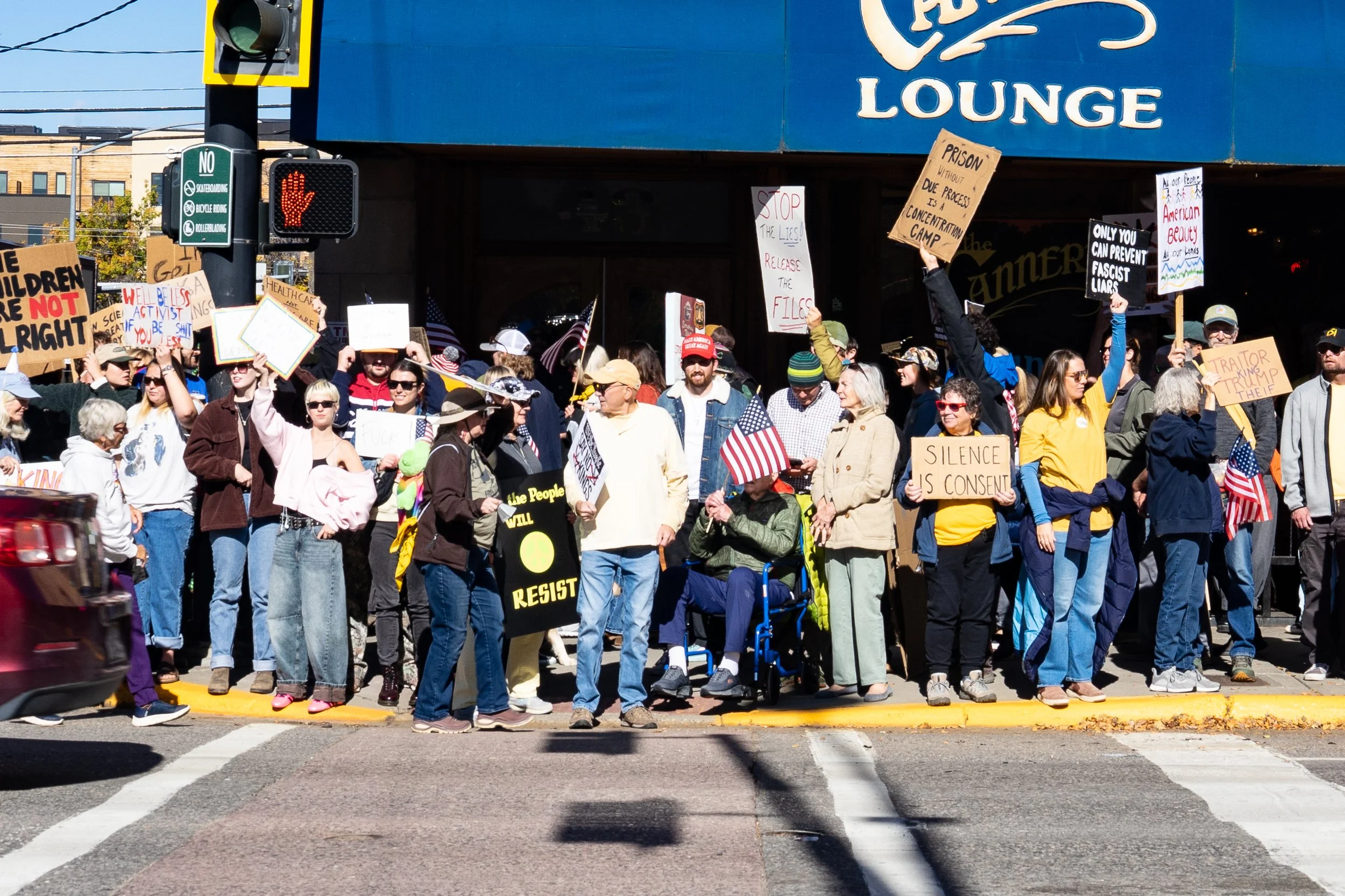 A crowd of protesters gathered on a city street corner, holding signs and American flags. They are standing in front of a blue building with a sign that reads 'Lounge.' The signs include messages about resistance, freedom, and political issues. The g