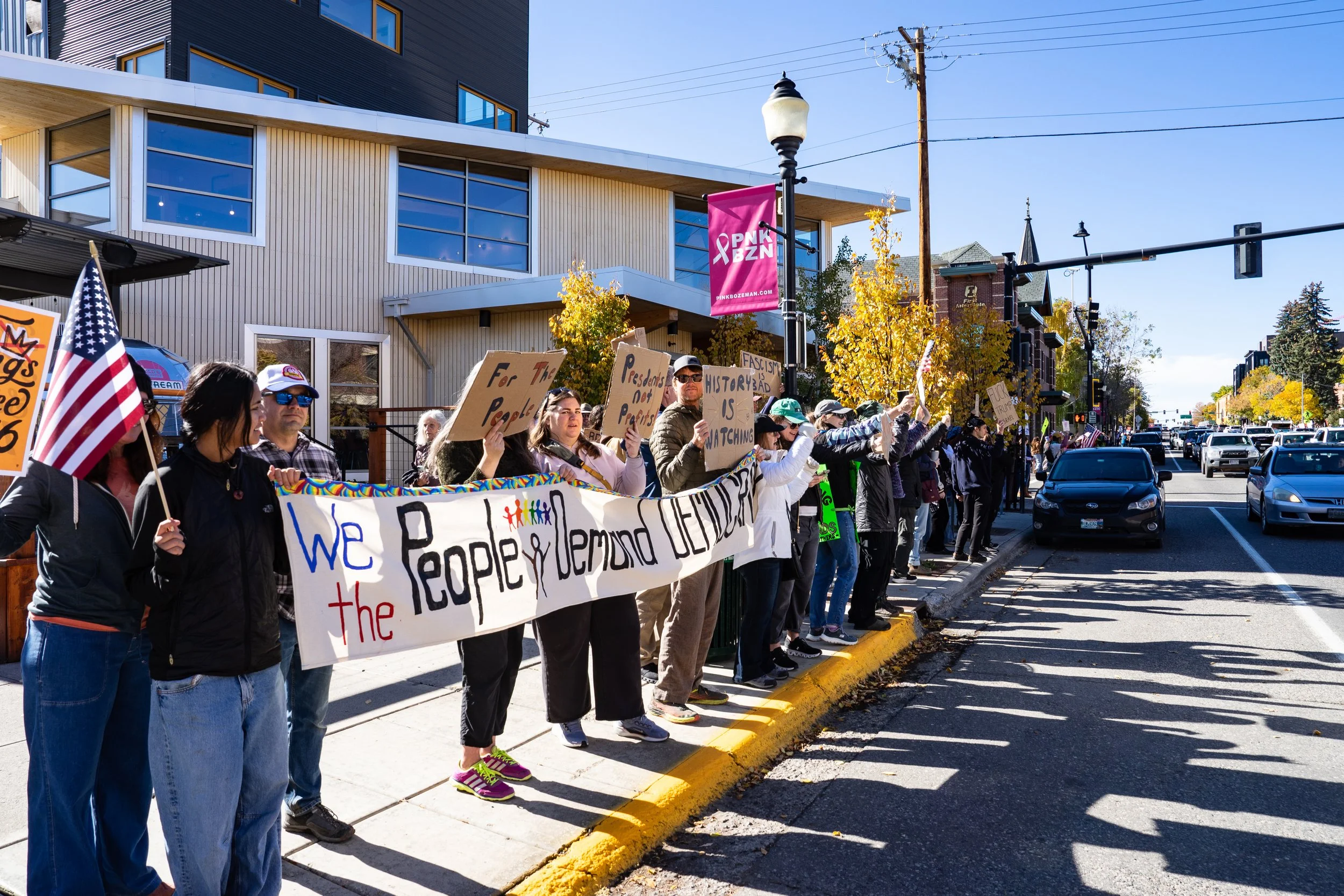 Group of people protesting on a city sidewalk, holding signs and a large banner that reads 'We the People Demand Democracy.' People are wearing casual attire with some holding flags. The street is busy with cars, and trees with autumn leaves line the