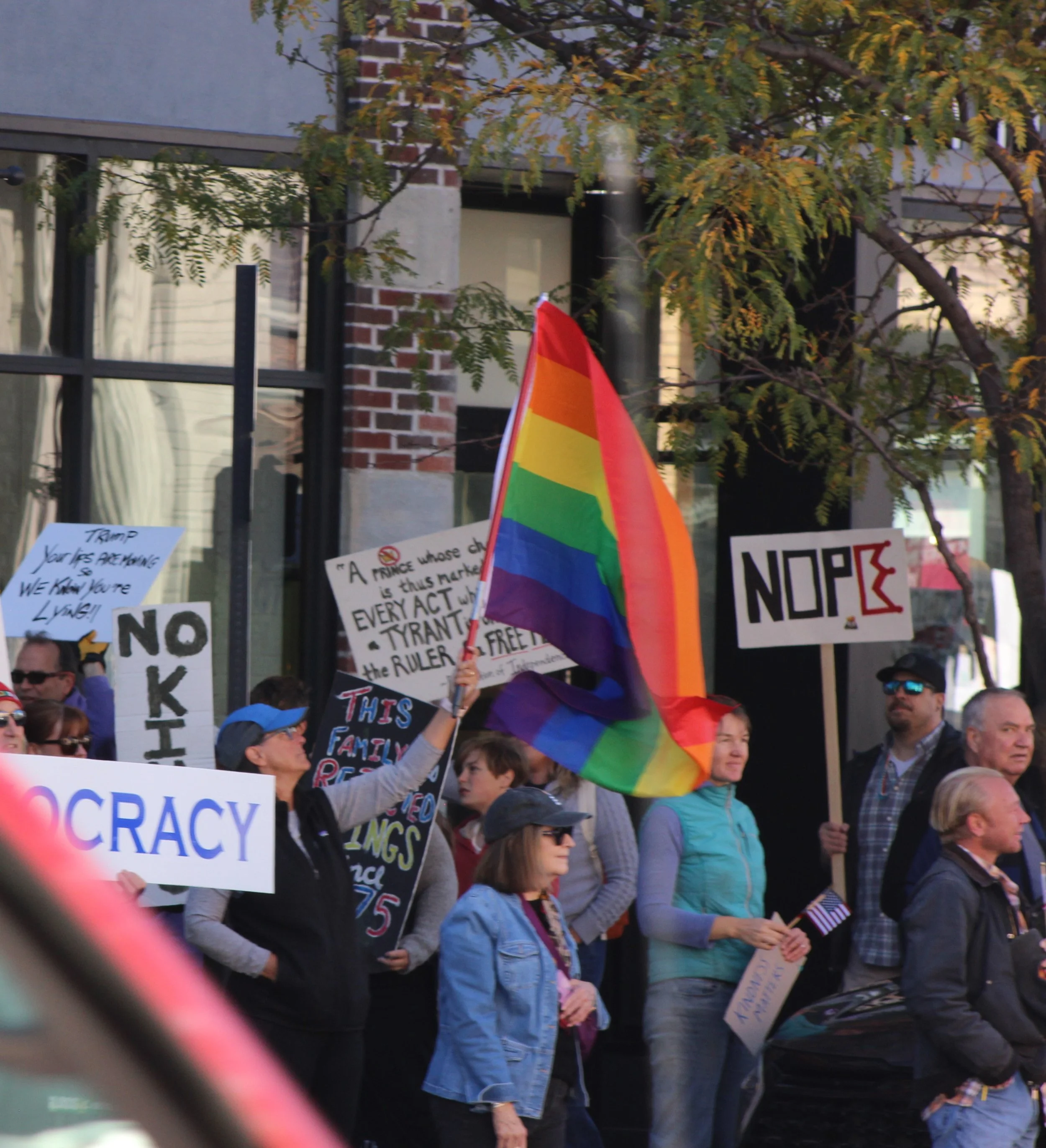 A crowd of people protesting with signs and a rainbow flag, standing outside a building with brick and glass windows, during daytime.