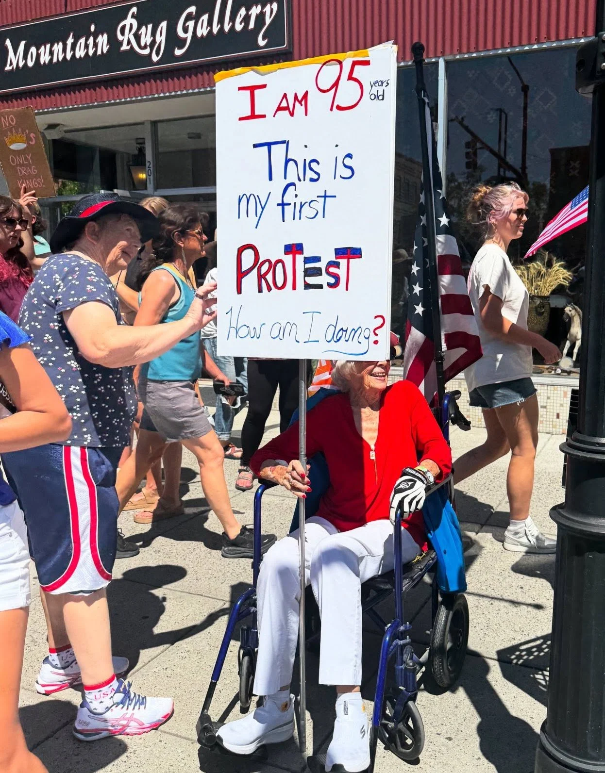 An elderly woman in a red jacket sitting in a wheelchair holding a sign that reads, 'I am 95 years old. This is my first protest. How am I doing?' surrounded by people at a protest or demonstration.