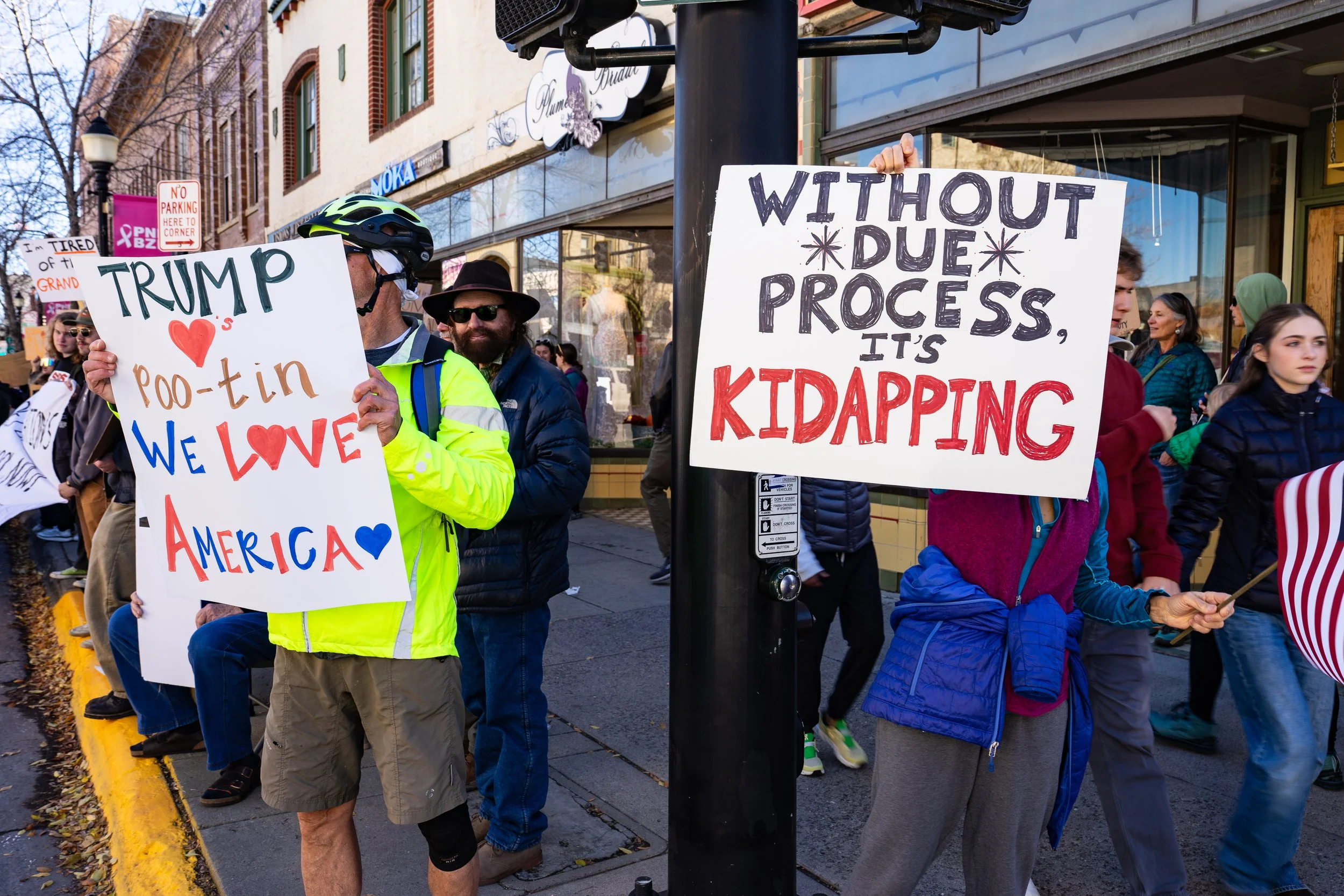 People participating in a protest march on a city street, holding signs with political messages. One sign reads 'TRUMP ♥️ roo-tin WE LOVE AMERICA,' and another sign reads 'WITHOUT DUE PROCESS, IT'S KIDNAPPING.' The crowd includes diverse individuals 