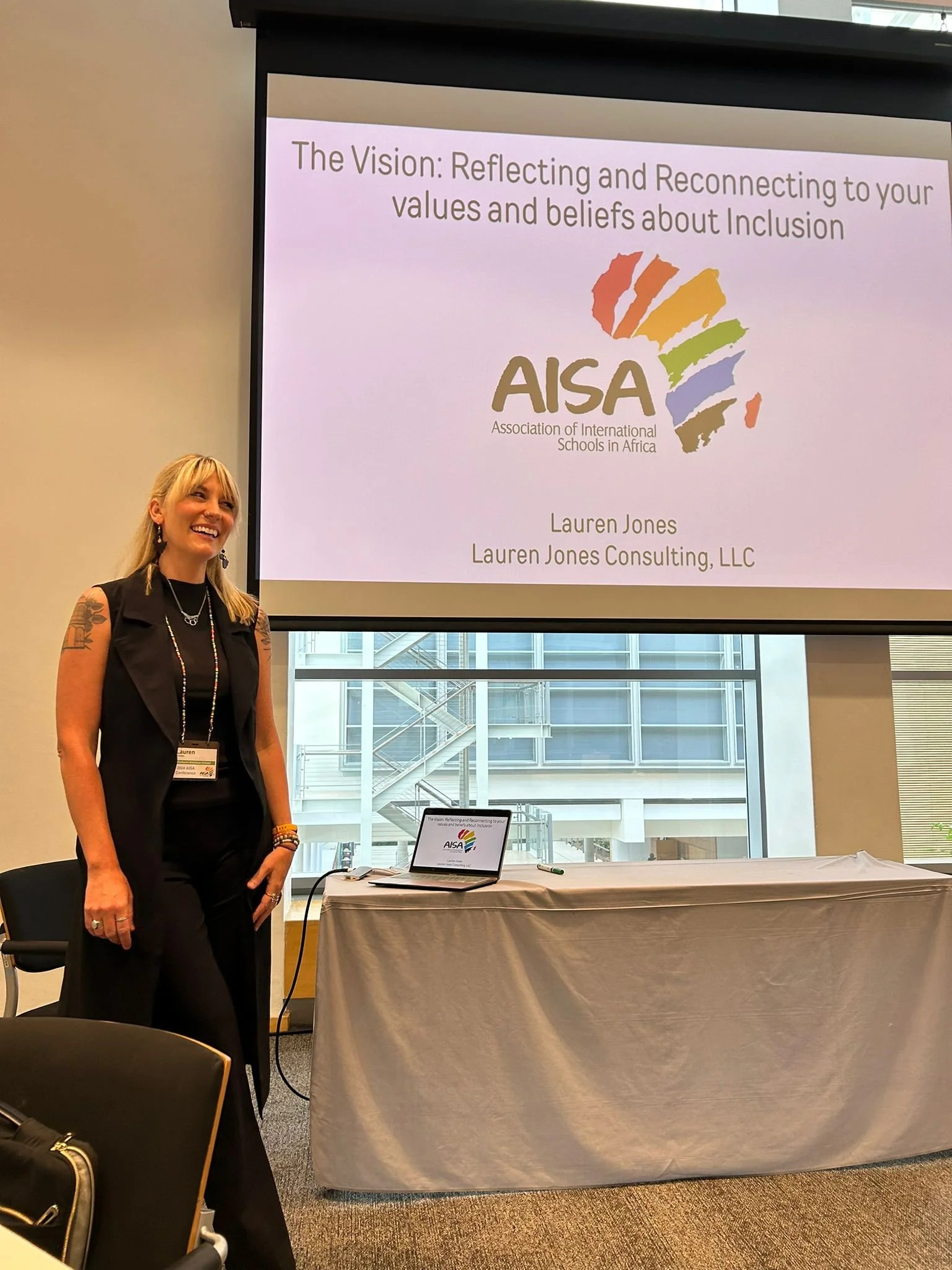 Woman standing beside a presentation screen showing a slide titled 'The Vision: Reflecting and Reconnecting to your values and beliefs about Inclusion'. The slide features a colorful logo of Africa and the text 'AISA, Association of International Schools in Africa', along with the name 'Lauren Jones' and 'Lauren Jones Consulting, LLC'. The woman has blonde hair, tattoos on her arms, and is wearing a black sleeveless outfit, jewelry, and a badge.