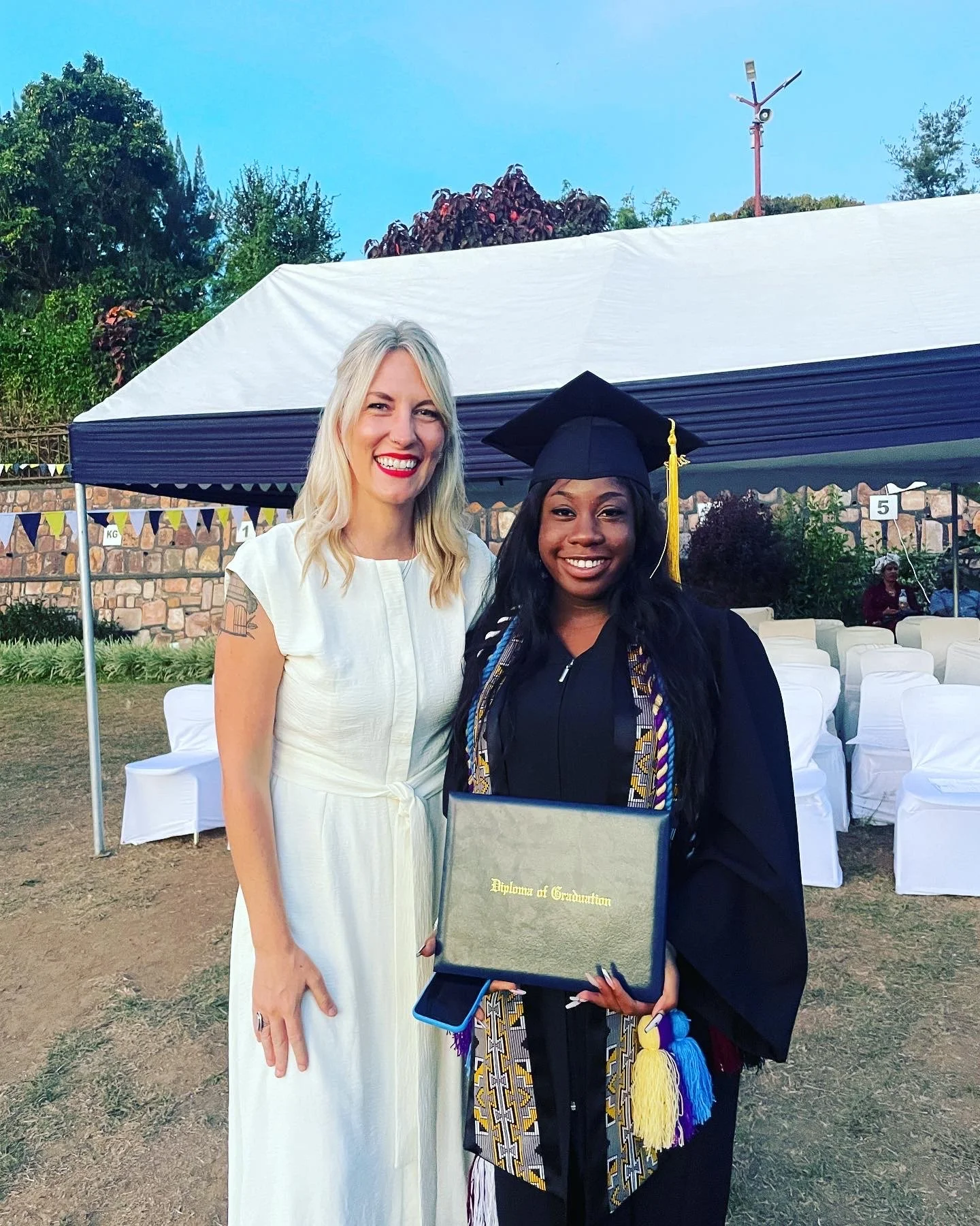 A young woman in a black graduation gown and cap holding a diploma stands next to an older woman in a white dress at an outdoor graduation ceremony. The young woman is smiling and the older woman is also smiling.