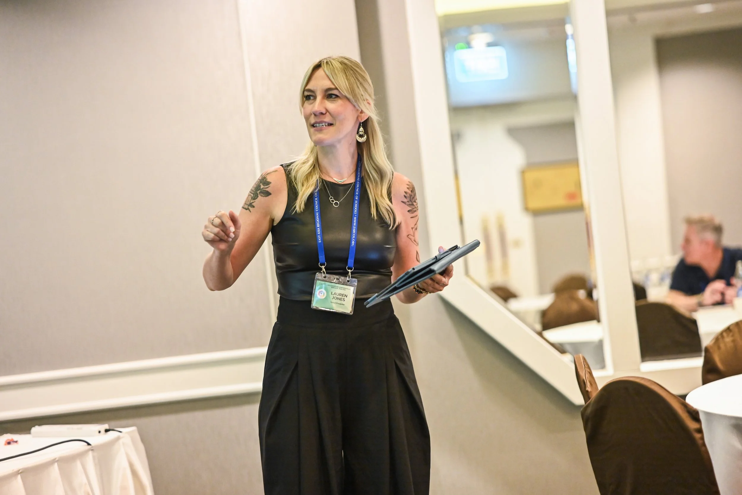 A woman with blonde hair, wearing a black dress and name badge, standing in a conference room, speaking or presenting.
