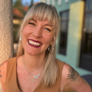 A smiling woman with blonde hair and bangs, wearing earrings, a necklace, and a sleeveless top, outdoors near a colorful building.