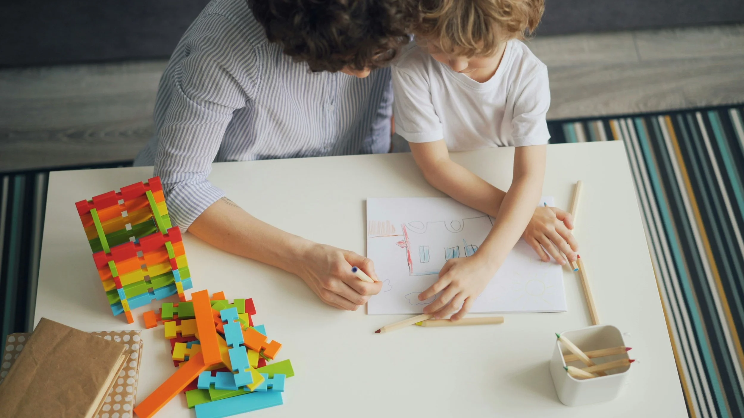 A woman and a young boy are sitting at a white table, drawing on paper with colored pencils. Colorful building blocks are stacked on the left side of the table, and a small container holds additional colored pencils on the right.
