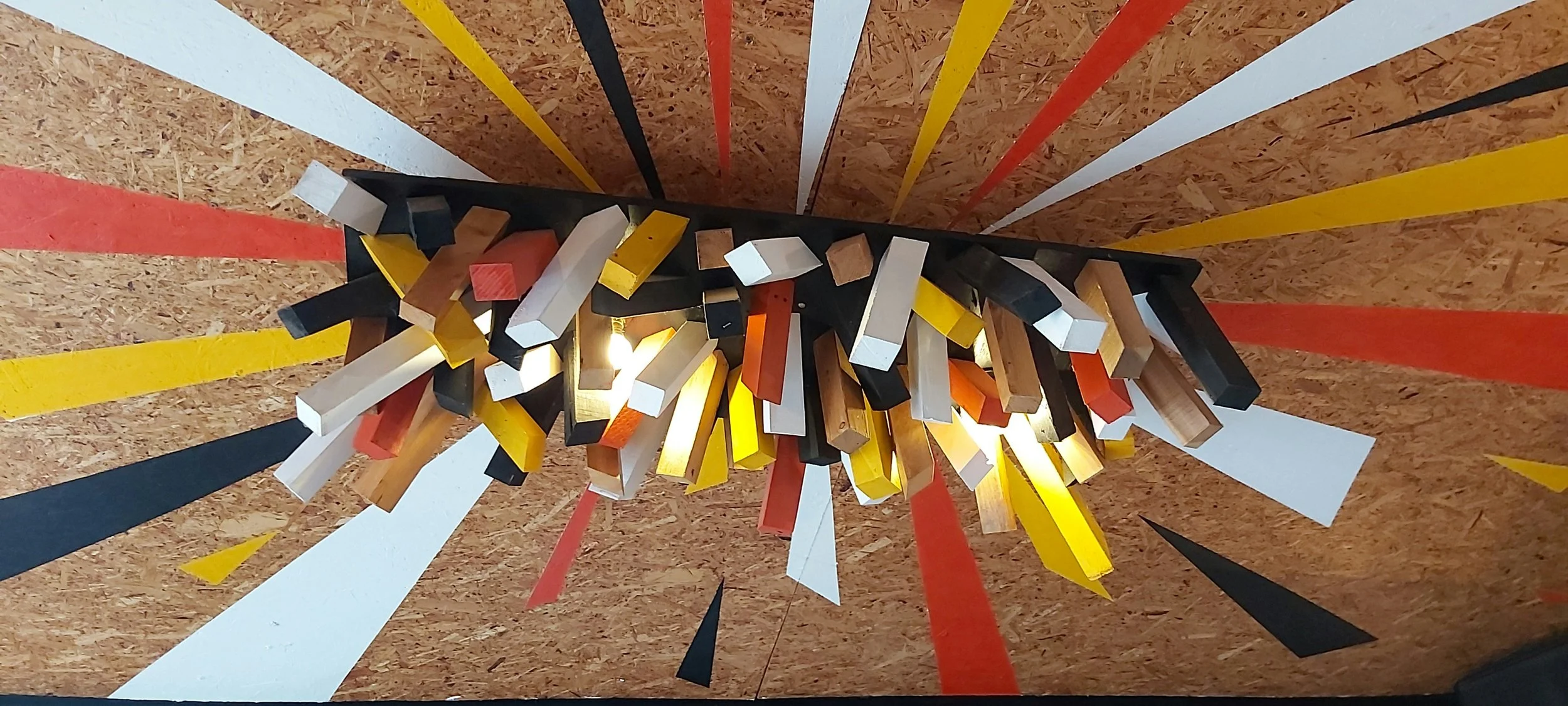 Colorful abstract chandelier with wooden and painted rectangular pieces hanging from black base on a textured wooden ceiling with painted lines.