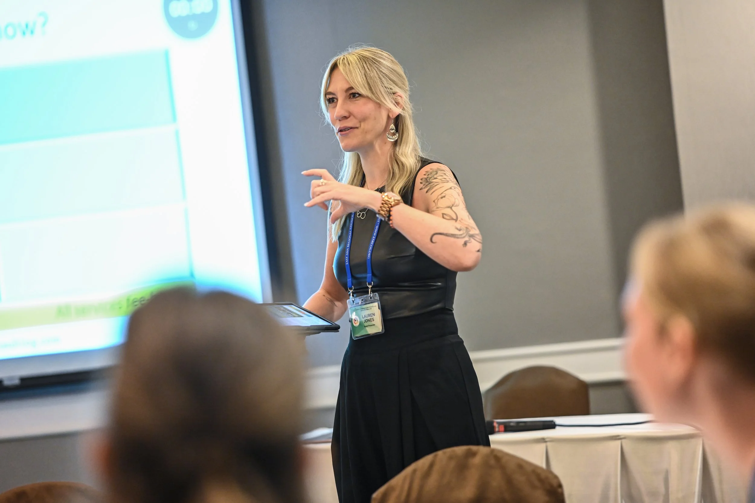 A woman with blonde hair, tattoos on her arms, wearing a black sleeveless top, giving a presentation in a conference room. She has a conference badge around her neck and is standing near a large screen, speaking to an audience.