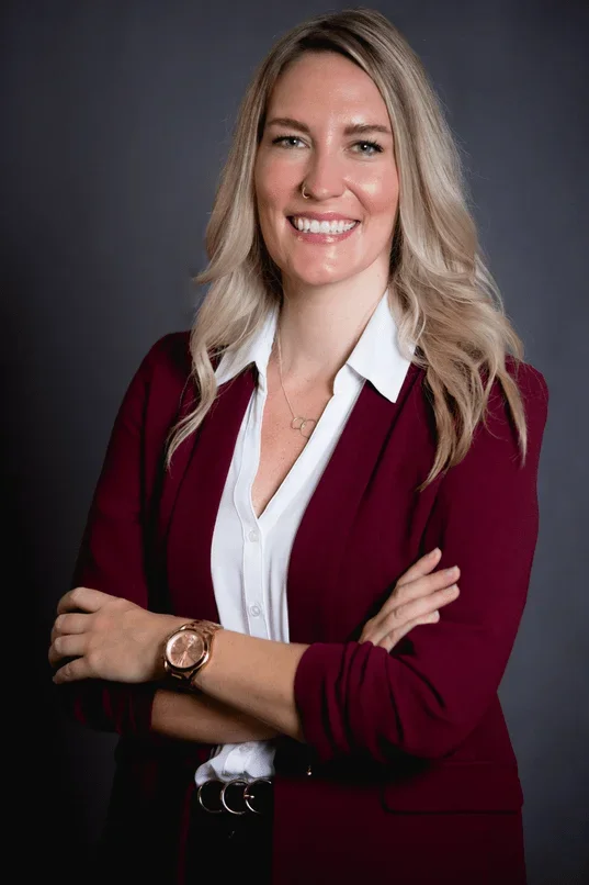 A woman with blonde hair, wearing a white shirt and burgundy blazer, smiling with arms crossed, against a dark background.