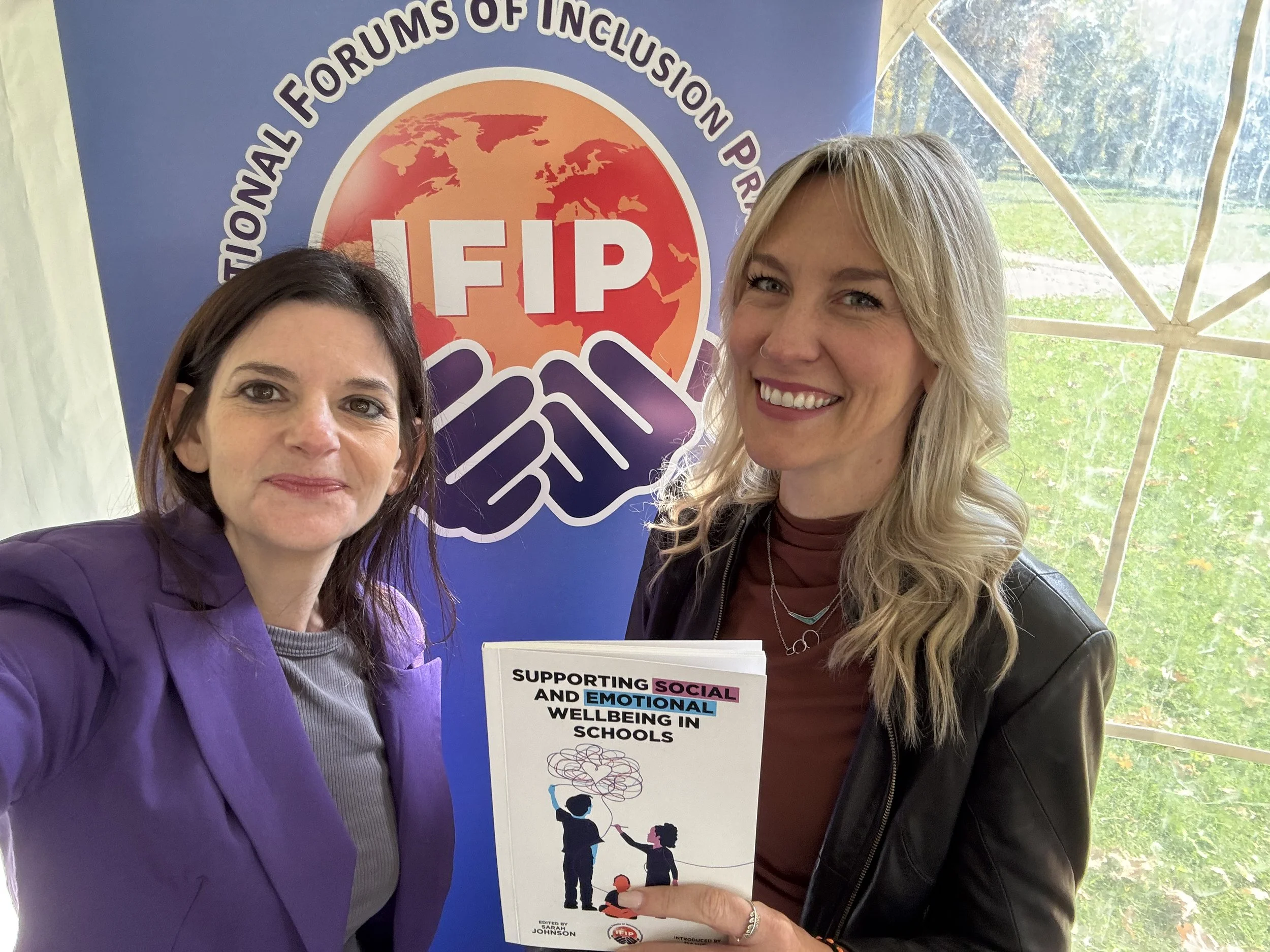 Two women smiling and holding a booklet titled "Supporting Social and Emotional Wellbeing in Schools" at an event for the International Forums of Inclusion Project, with a FIP banner in the background.