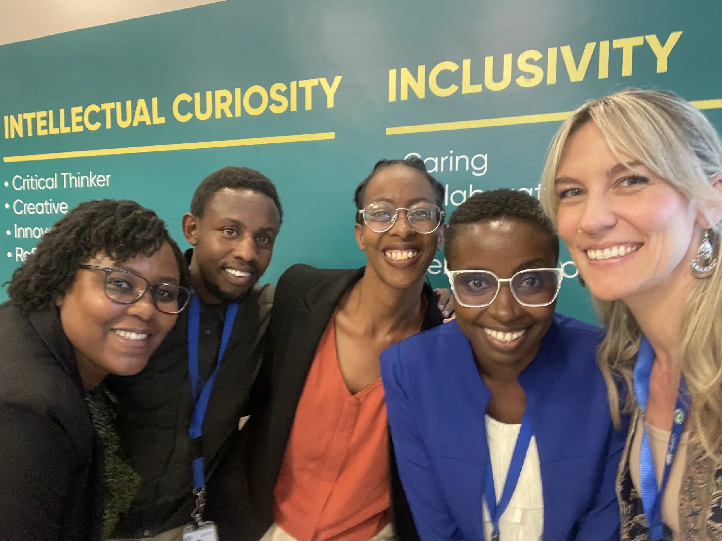A group of five smiling individuals taking a selfie in front of a blue-green background with words like 'Intellectual Curiosity' and 'Inclusivity'; they are wearing conference badges and professional attire.