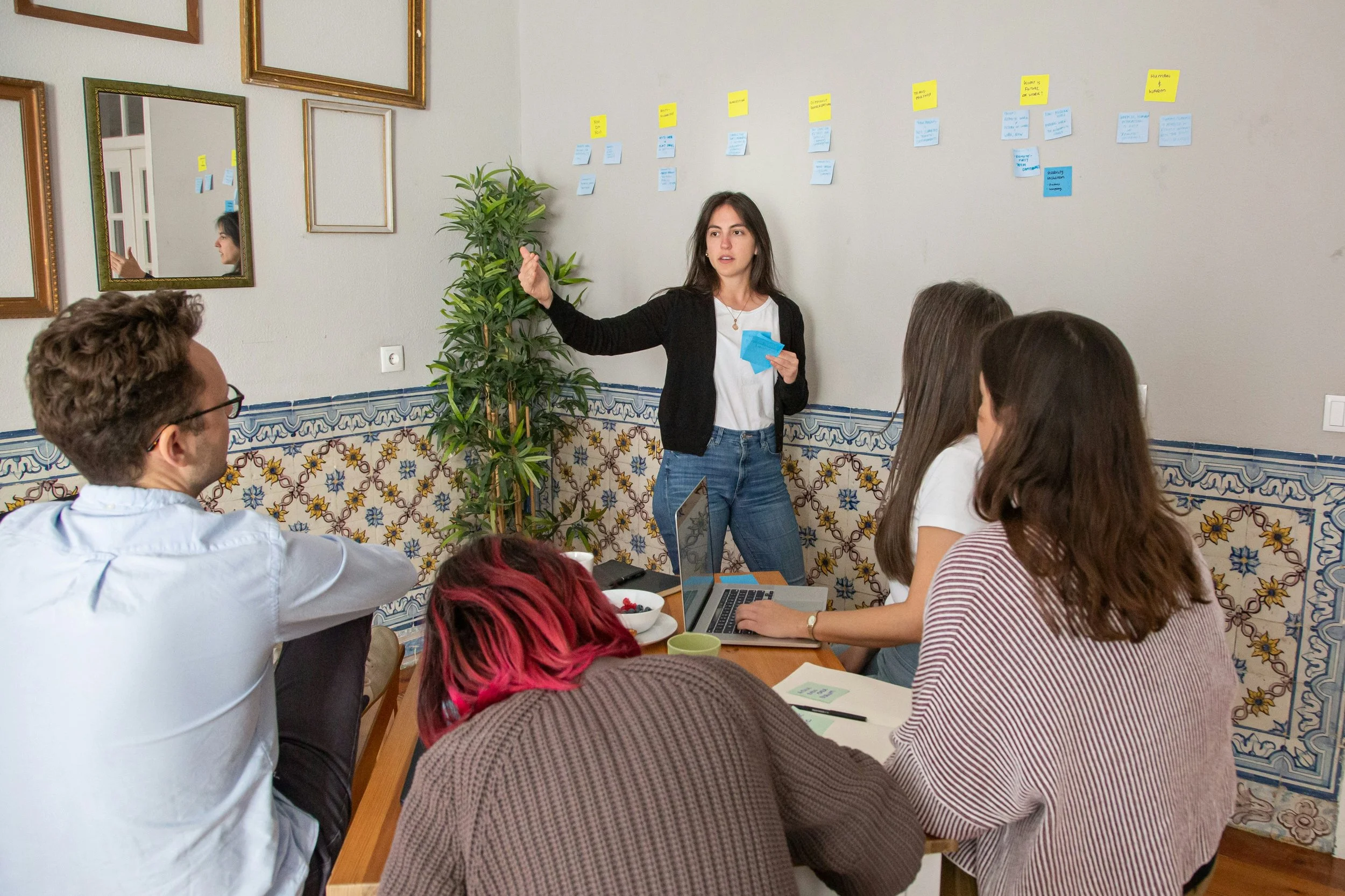A woman presenting to a group of four people seated around a table with laptops and notebooks, in a room decorated with framed pictures and blue patterned tiles, with sticky notes on the wall.