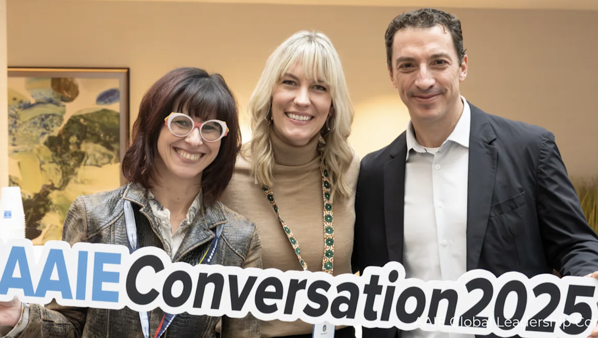 Three smiling women and one smiling man standing side by side, holding a sign that reads "AAIE Conversation 2025" at an indoor event.