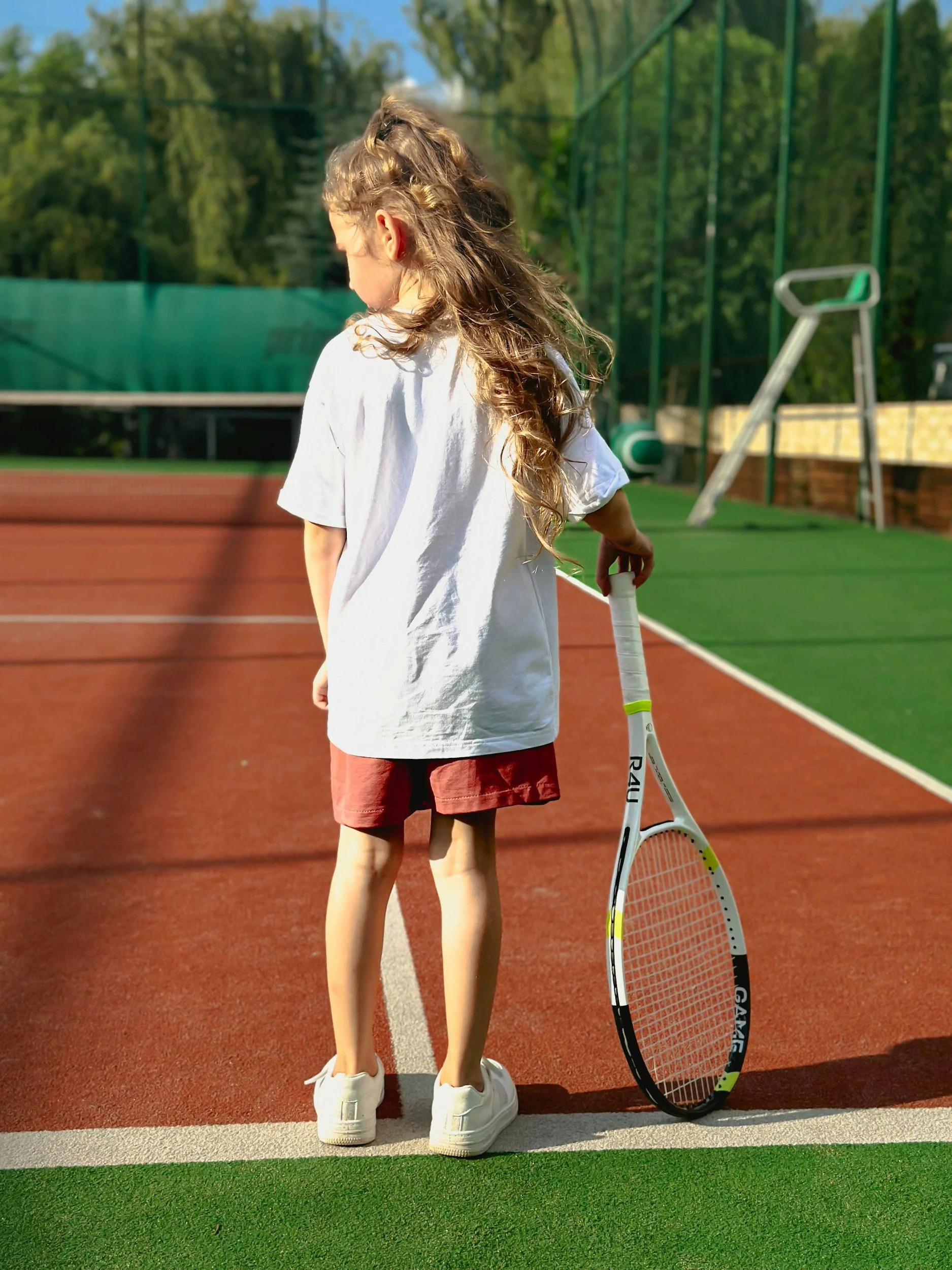 Young girl standing on a tennis court holding a tennis racket, with trees and a green fence in the background.