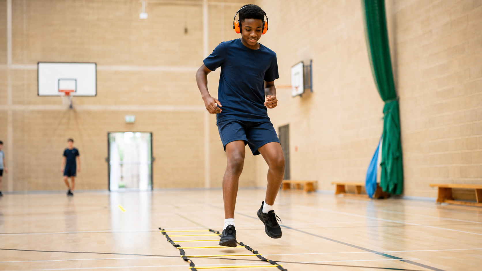 Boy in navy sportswear wearing orange headphones doing agility ladder exercises in a gymnasium with basketball hoops and other children in the background.