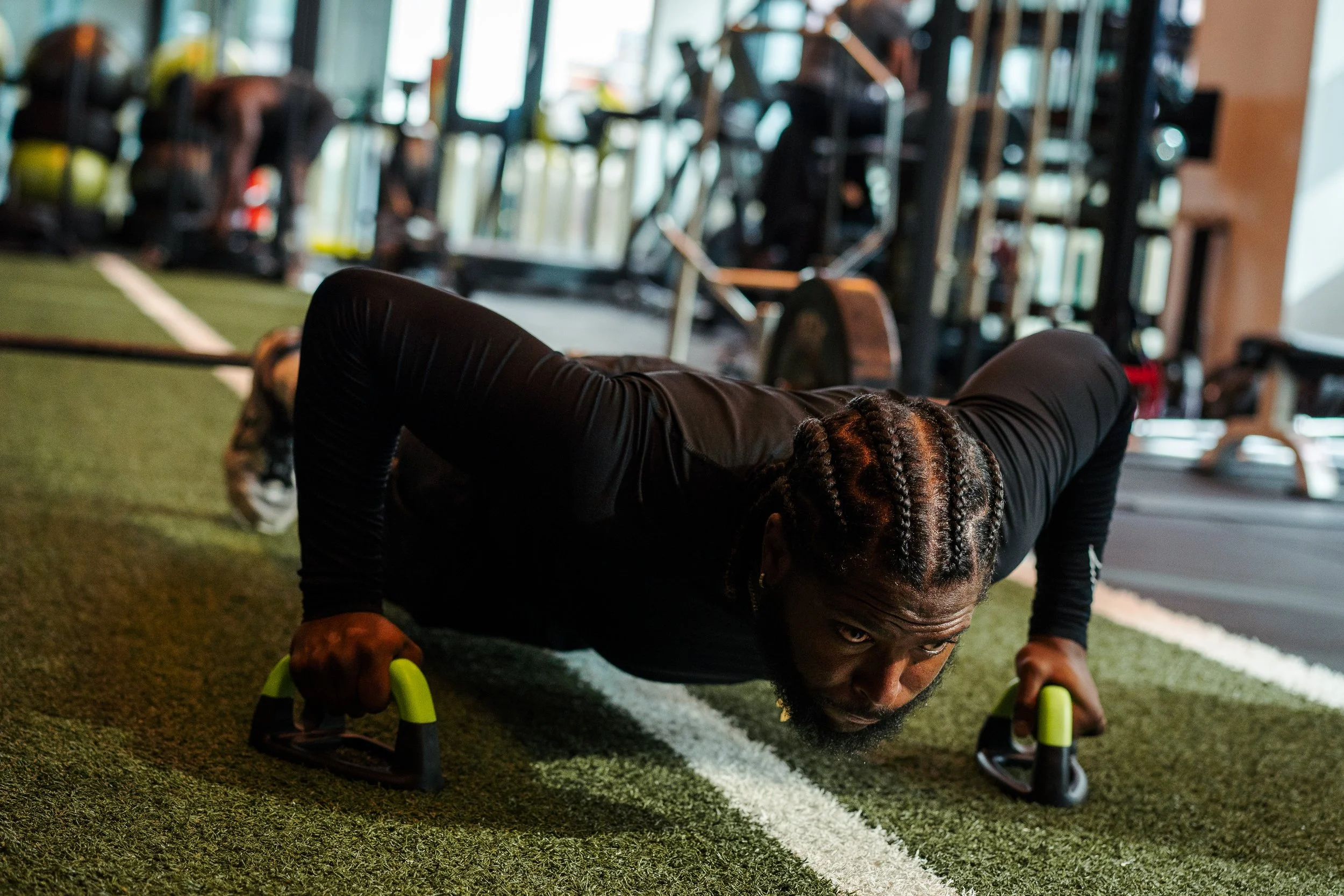 A man with braided hair doing push-ups on a green turf with white lines in a gym.