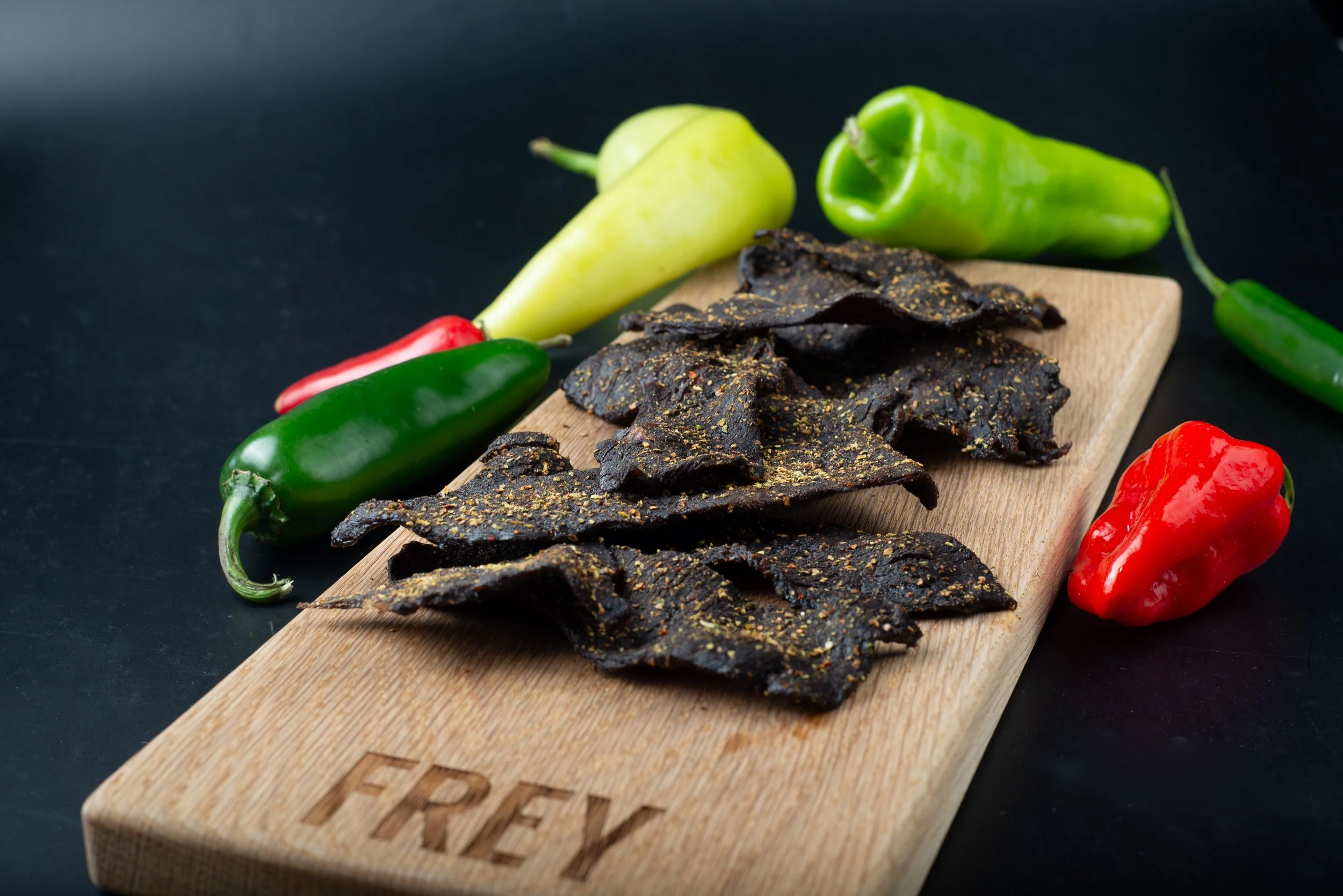 A wooden cutting board with the word 'FREY' engraved, holding several dark, seasoned beef jerky slices. Around the board are whole green, red, and yellow peppers, and a green chili pepper on a dark background.