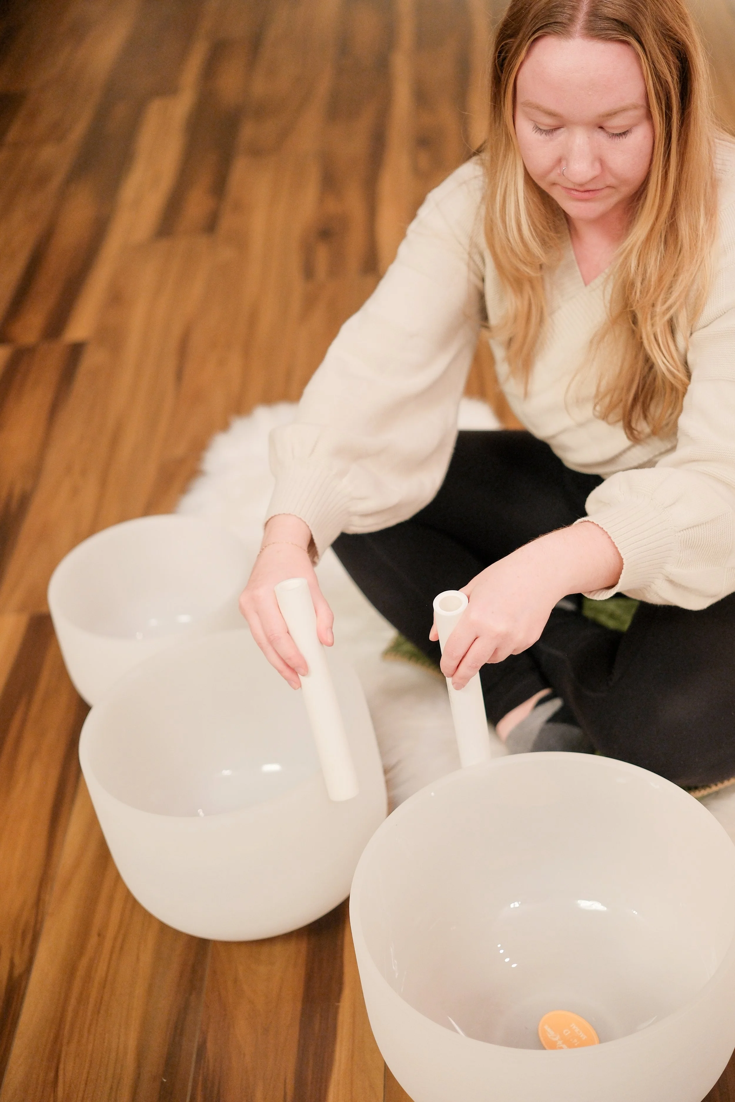 A woman sitting on the floor with three white crystal singing bowls in front of her, holding a mallet in each hand.