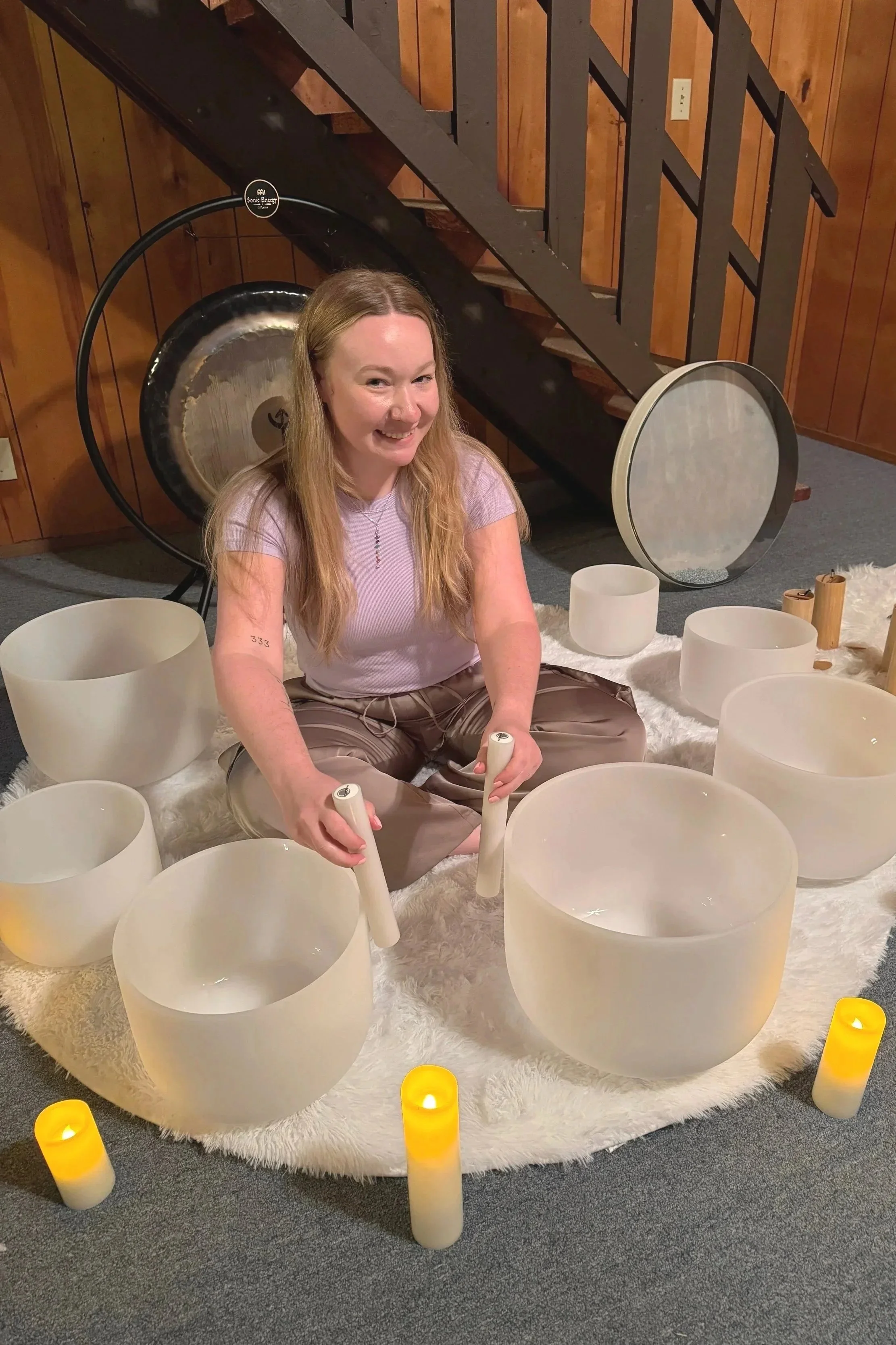 A woman with long blonde hair sitting on a white furry rug surrounded by large white crystal singing bowls, holding mallets, with a gong and wooden percussion instruments in the background, lit by candles.