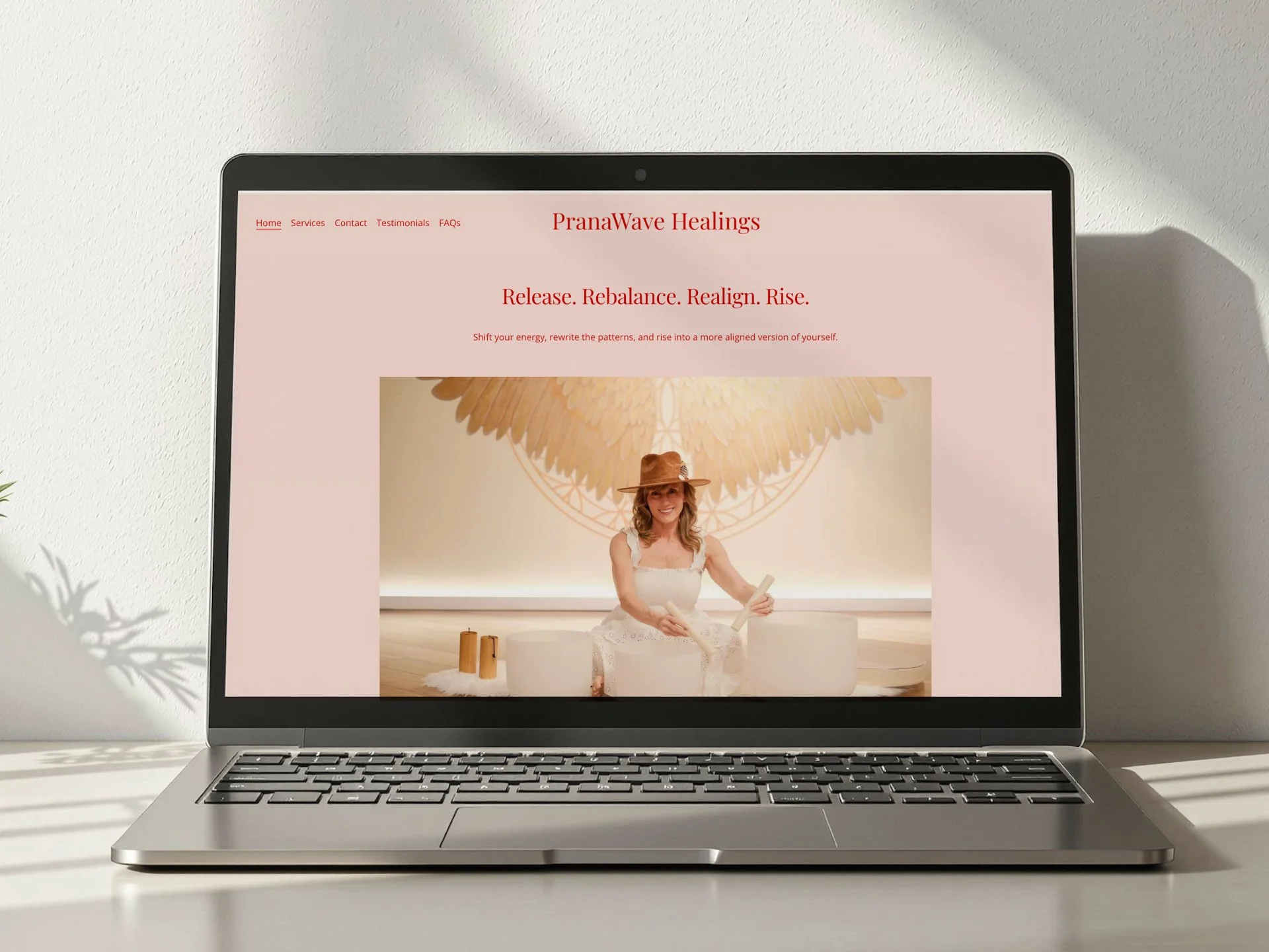 Laptop screen displaying a website for PranaWave Healings with a woman sitting cross-legged, smiling, in a white dress and floppy hat, holding drums, with a large feather mural behind her.