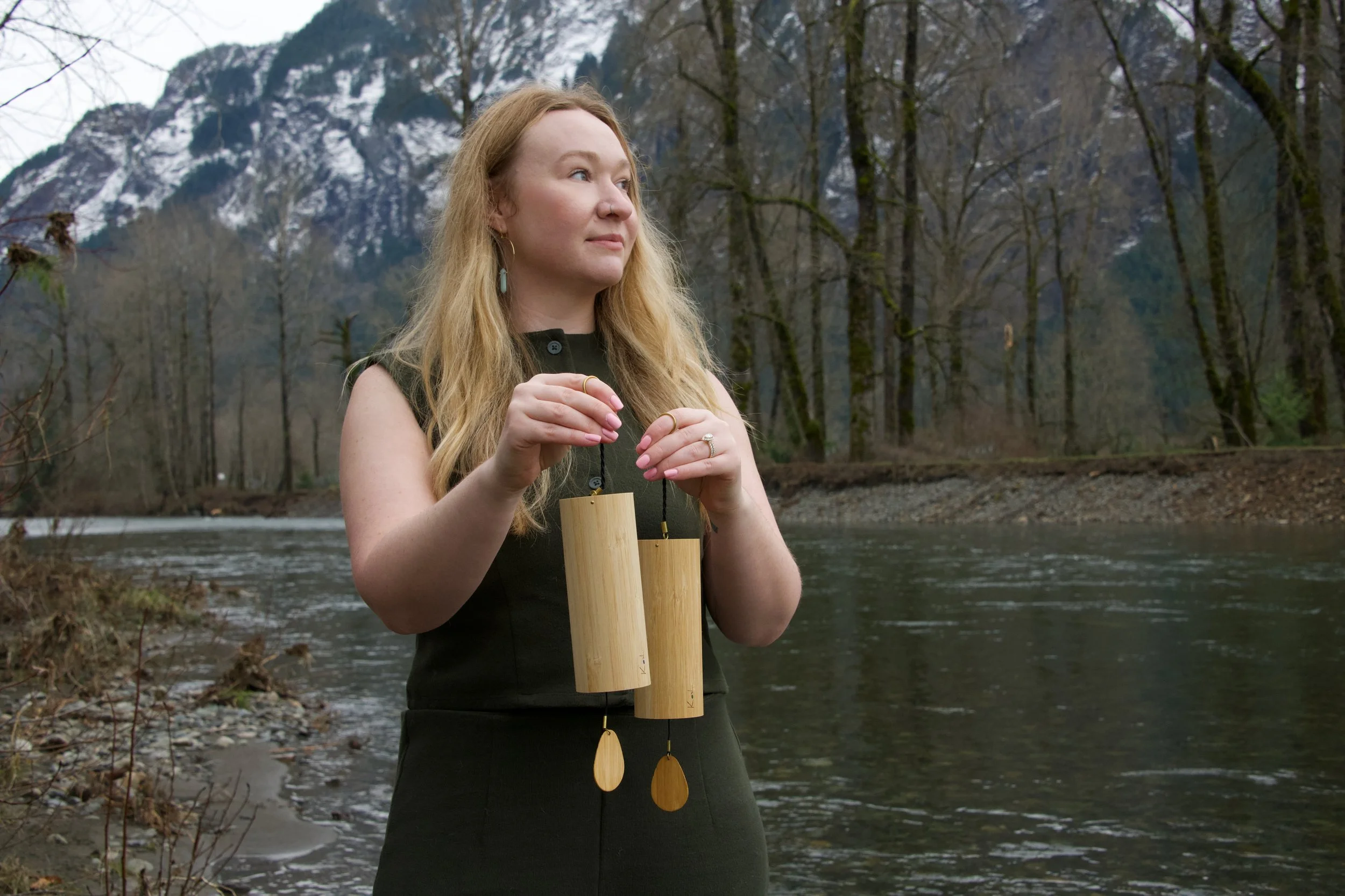 A woman standing near a river holding two large wooden wind chimes, with mountains and bare trees in the background.