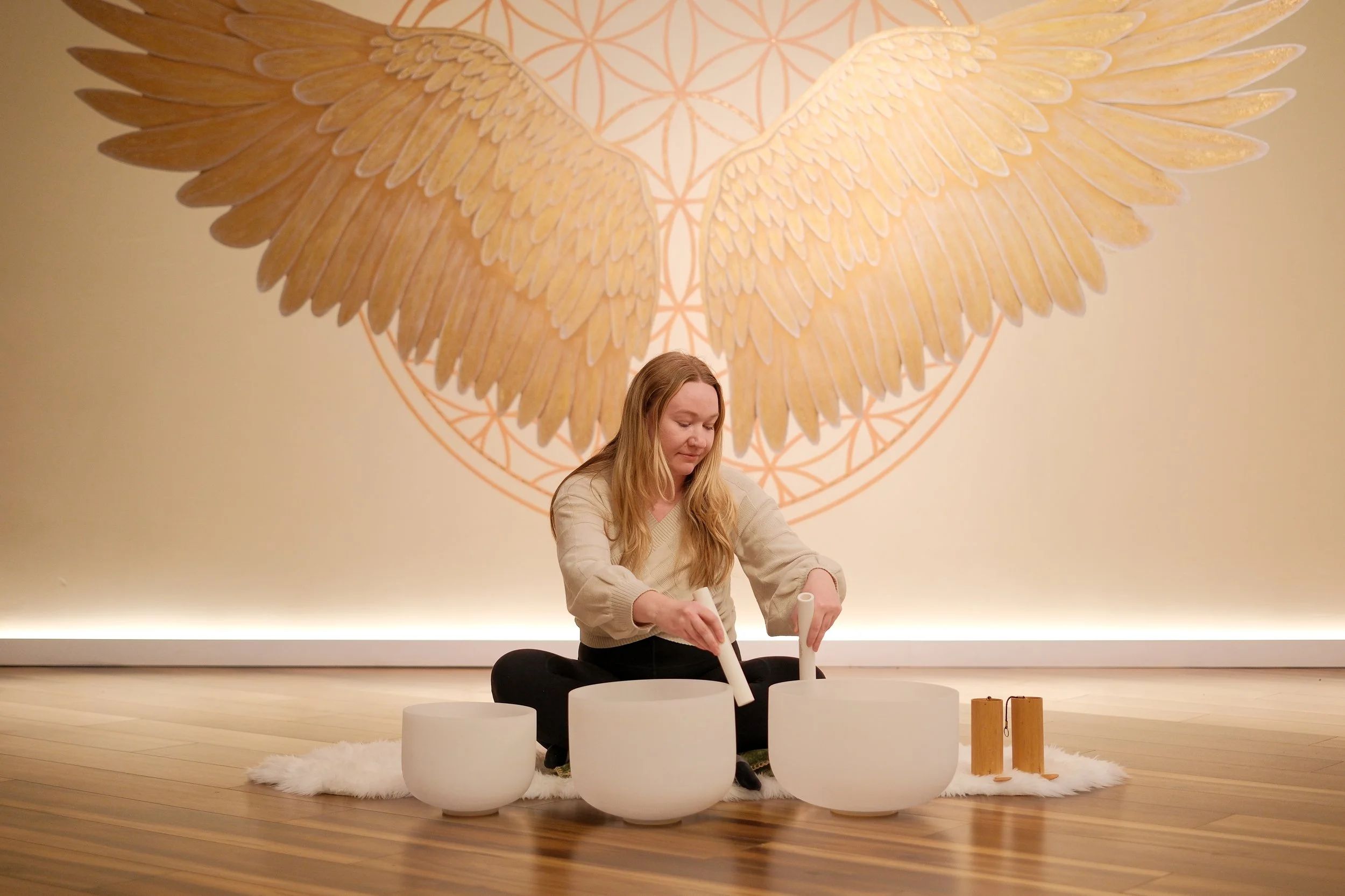 A woman sitting on a white furry rug playing crystal singing bowls in front of a large wall mural of golden angel wings and geometric patterns.