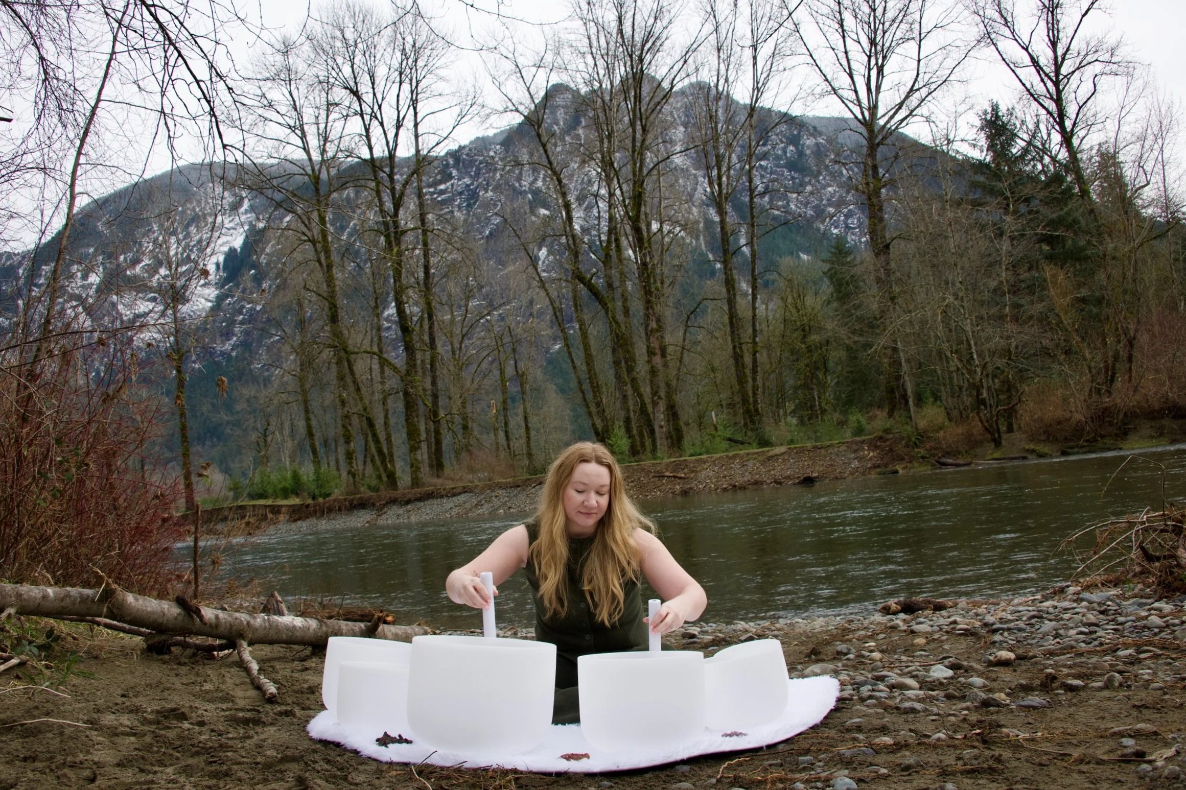 A woman with long blonde hair is playing crystal bowls by a river in a forested mountain area.