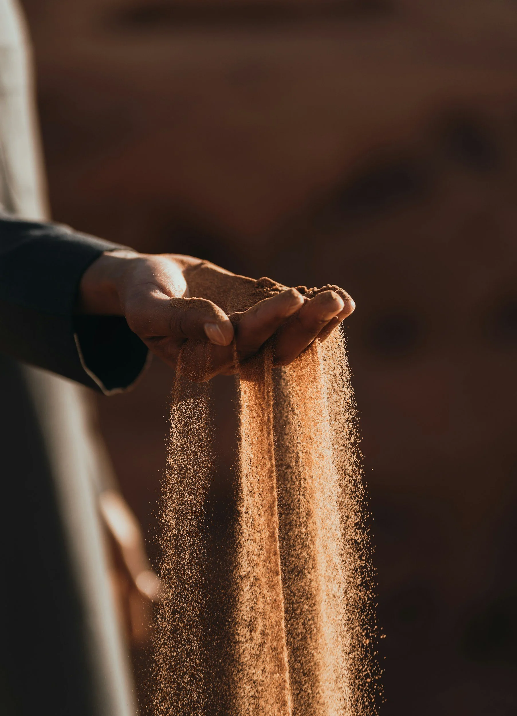 Close-up of a hand holding and pouring fine, reddish-brown sand against a blurred background.