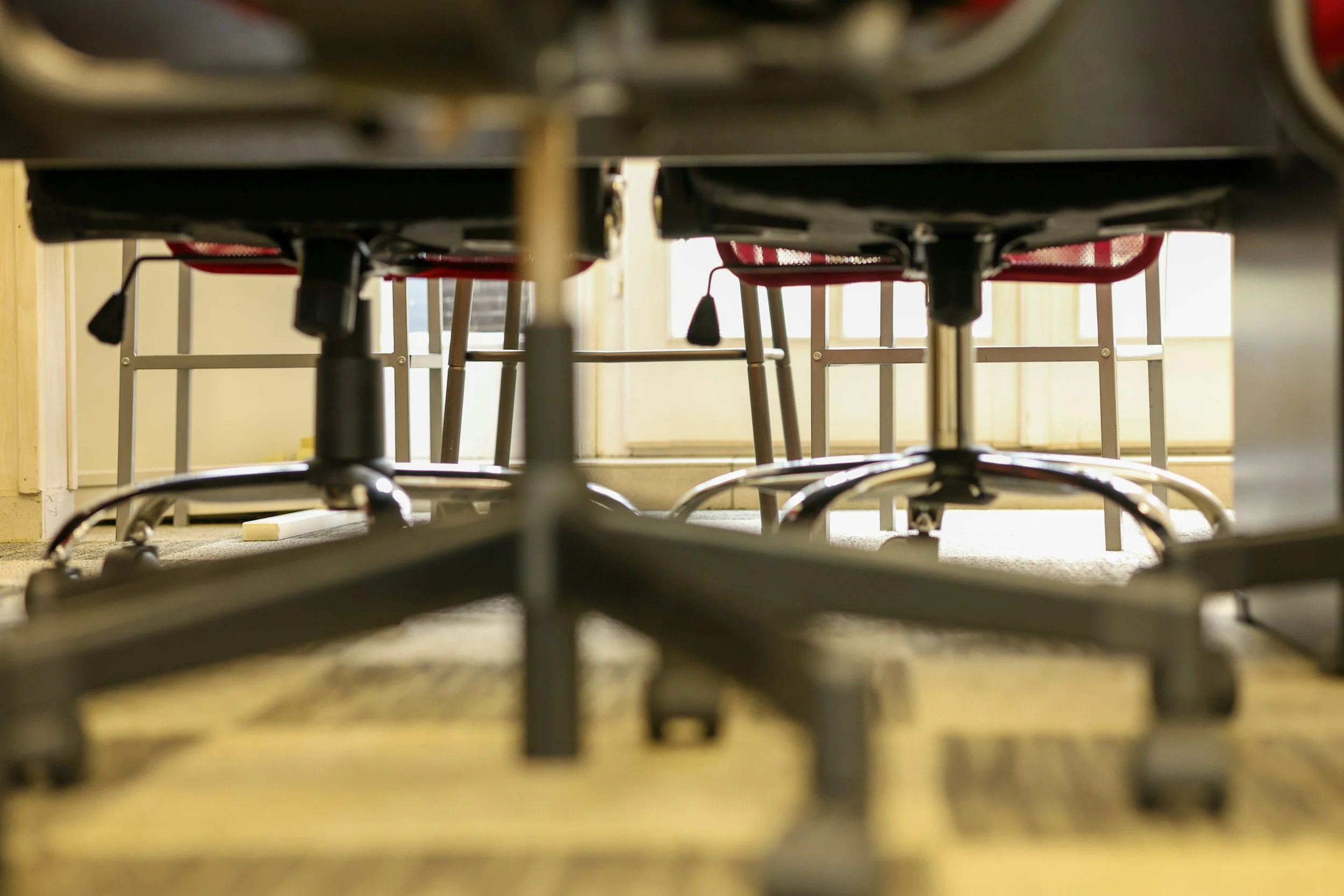 View from underneath tables and chairs in a classroom or cafeteria, looking through the legs at the floor and bright windows in the background.
