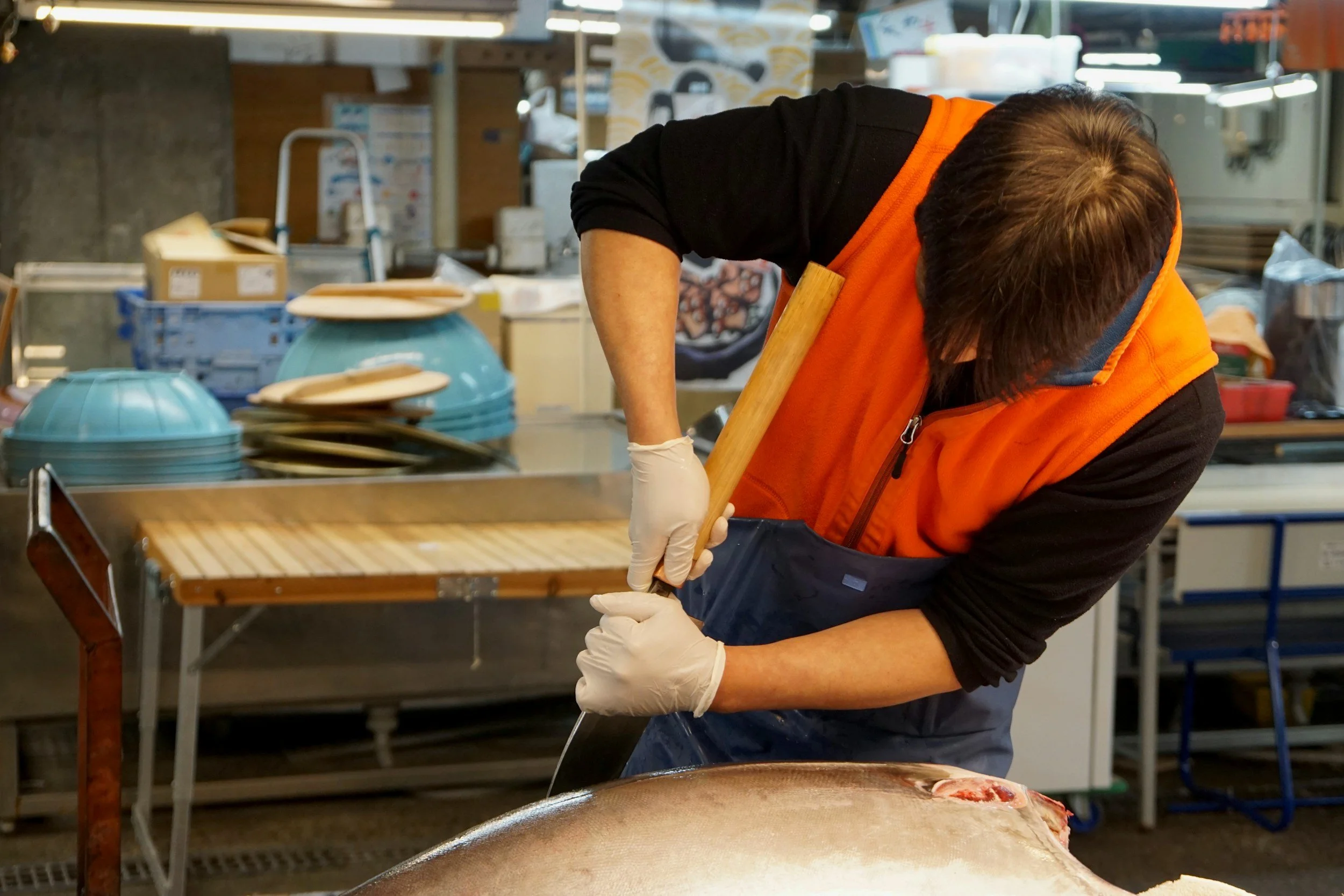 A person in an orange vest and gloves is filleting a fish at a seafood market or kitchen, with various dishes and kitchen equipment in the background.