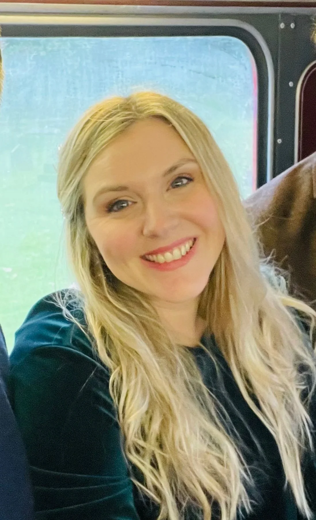 A woman with long blonde hair smiling, wearing a dark green or black velvet top, on a bus or train with a window showing greenery outside.