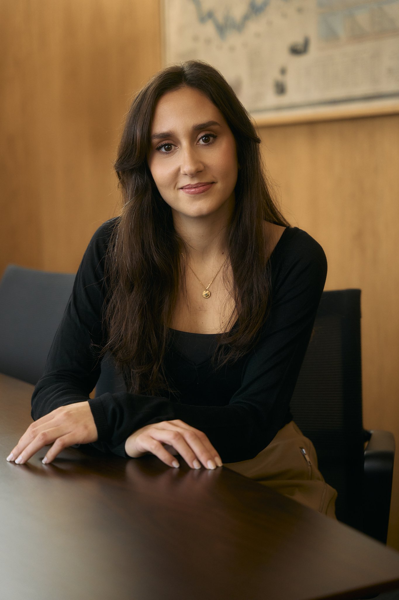 Mulher com cabelo castanho escuro, vestindo blusa preta e saia, sentada em uma mesa de madeira, com parede de madeira ao fundo e quadro pendurado na parede.