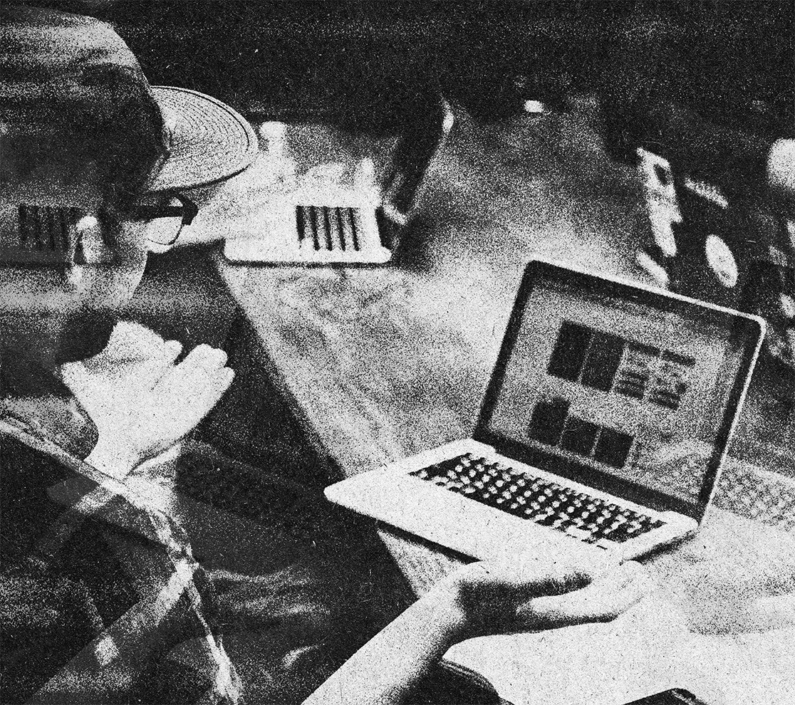 A person wearing a baseball cap and glasses sitting at a desk, working on a laptop, with a notebook and other objects on the desk.