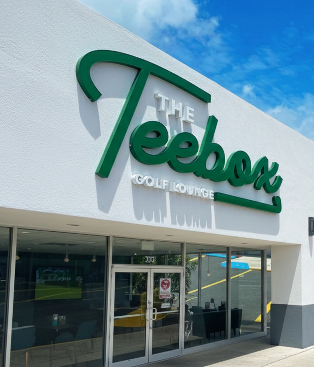 Exterior of The Teebox Golf Lounge building with a large sign featuring green lettering and glass doors, under a blue sky with clouds.
