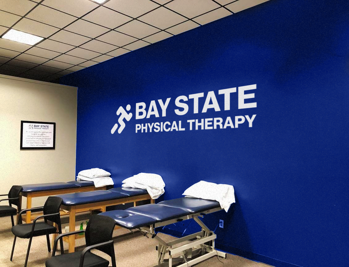 A physical therapy clinic room with two therapy tables covered with white towels, chairs, and a sign on the wall stating "Bay State Physical Therapy" and its logo.