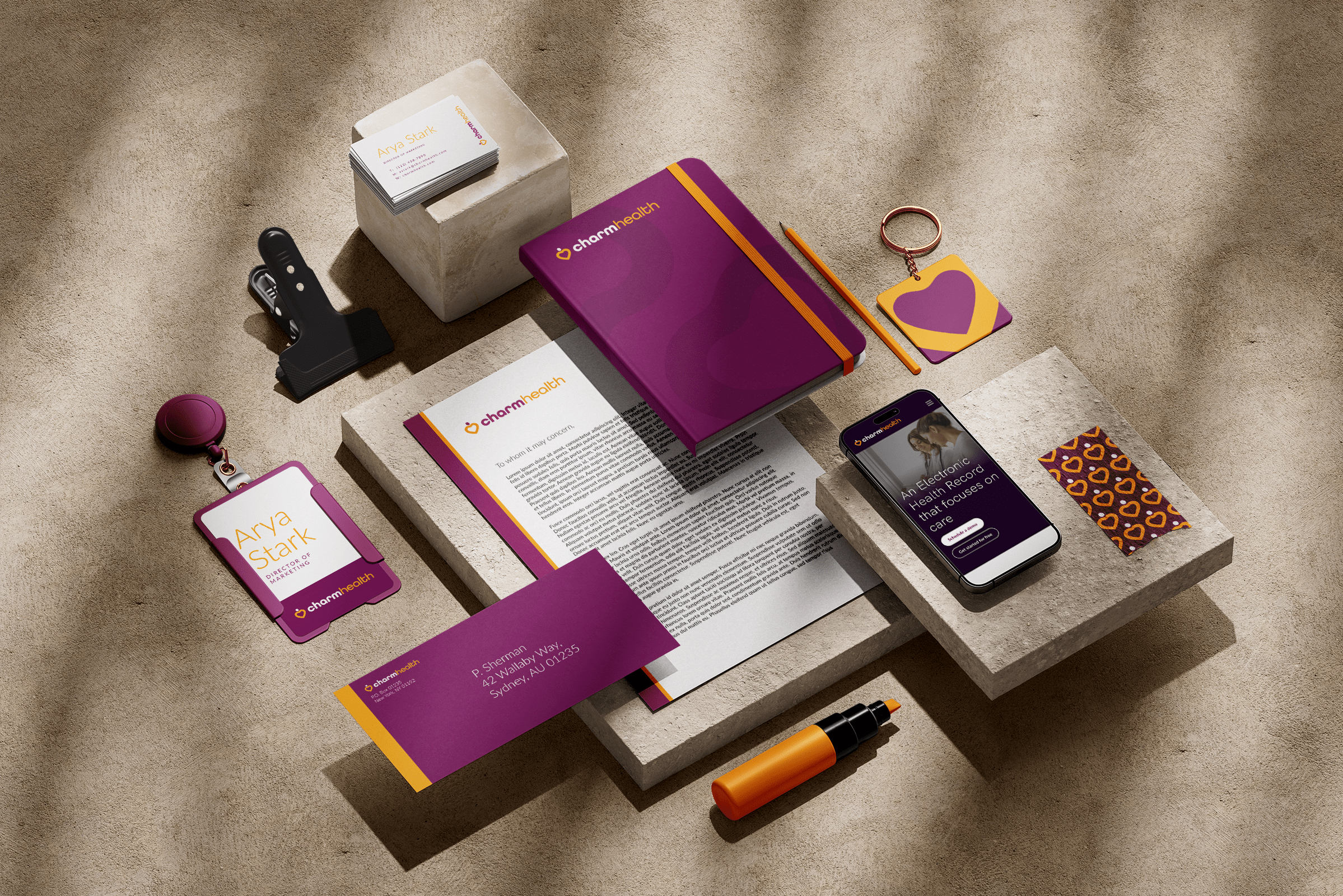 Collection of Charmhealth branded office items including a purple notebook, printed documents, a smartphone displaying a Charmhealth app, business cards, a badge with the name Arya Stark, a keychain, and various office accessories on concrete surfaces.
