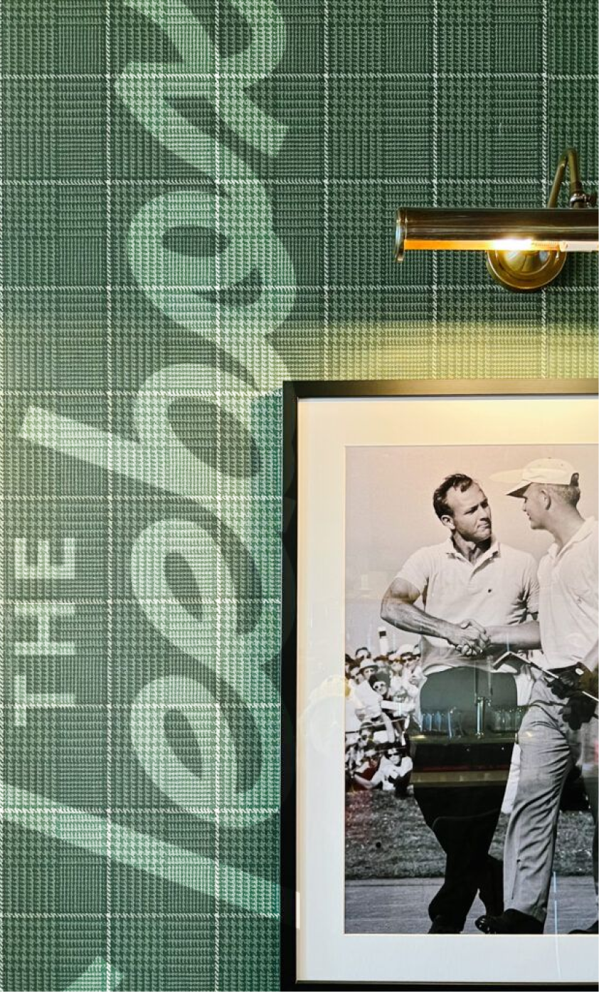 Part of an interior wall with green plaid patterned wallpaper and a black and white framed photograph of two men shaking hands at a golf course, with one wearing a cap and the other not, beneath a mounted wall light. The wallpaper features a subtle floral design.
