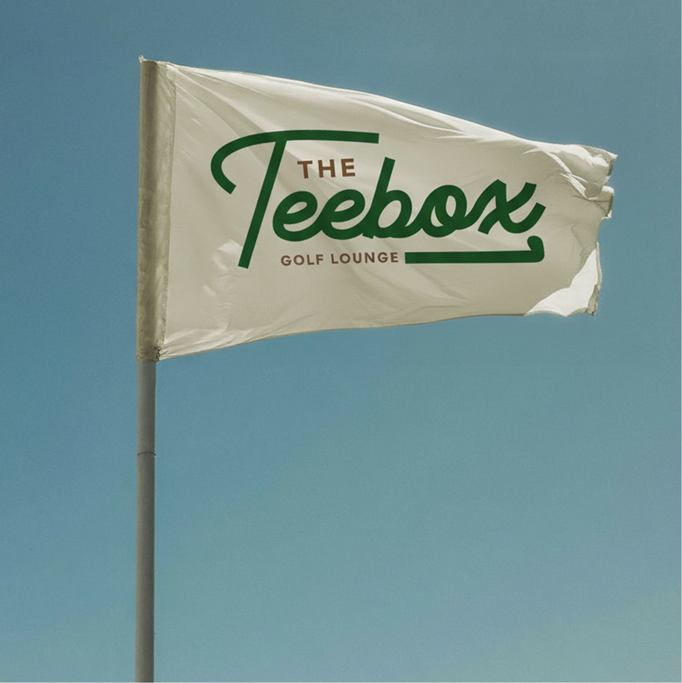 A white flag with green and brown text reading 'The Teebox Golf Lounge' is flying against a clear blue sky.